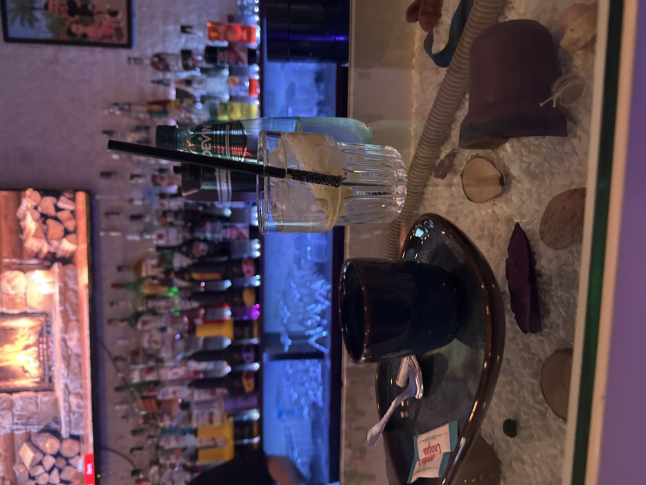 The image depicts an indoor bar or lounge environment located in Varna, Bulgaria. In the foreground, a glass-topped table features a decorative base filled with white crushed stone. Scattered across this base are natural elements, including dried leaves (one deep purple, others brown), light-colored wood slices, several peanuts or small nuts, a coiled light-brown tube (possibly a hookah hose), and a dark, irregularly shaped object appearing to be ceramic or stone. On the glass surface of the table, a dark brown ceramic cup rests on a matching irregular-shaped saucer, accompanied by a small metallic spoon and a white packet labeled "Lavazza." Adjacent to these items, a clear, ribbed glass contains a beverage with ice cubes and lemon slices, fitted with a black drinking straw. Directly behind this beverage glass, an upright plastic bottle of "Devin" mineral water is visible.
The midground reveals a long bar counter illuminated by prominent blue LED underlighting. Numerous bottles of various alcoholic beverages are arranged on shelves behind the counter. A partial view of a person's upper left arm and shoulder is visible on the far left, indicating the presence of a bartender or staff member.
The background features a large flat-screen display mounted above the bar, depicting a digital image of a burning fireplace with stacked logs. To the right of this screen, a framed picture or artwork with tropical imagery, including a palm tree and indistinct figures, is visible. The wall surface behind the bar and display exhibits a textured or patterned finish.