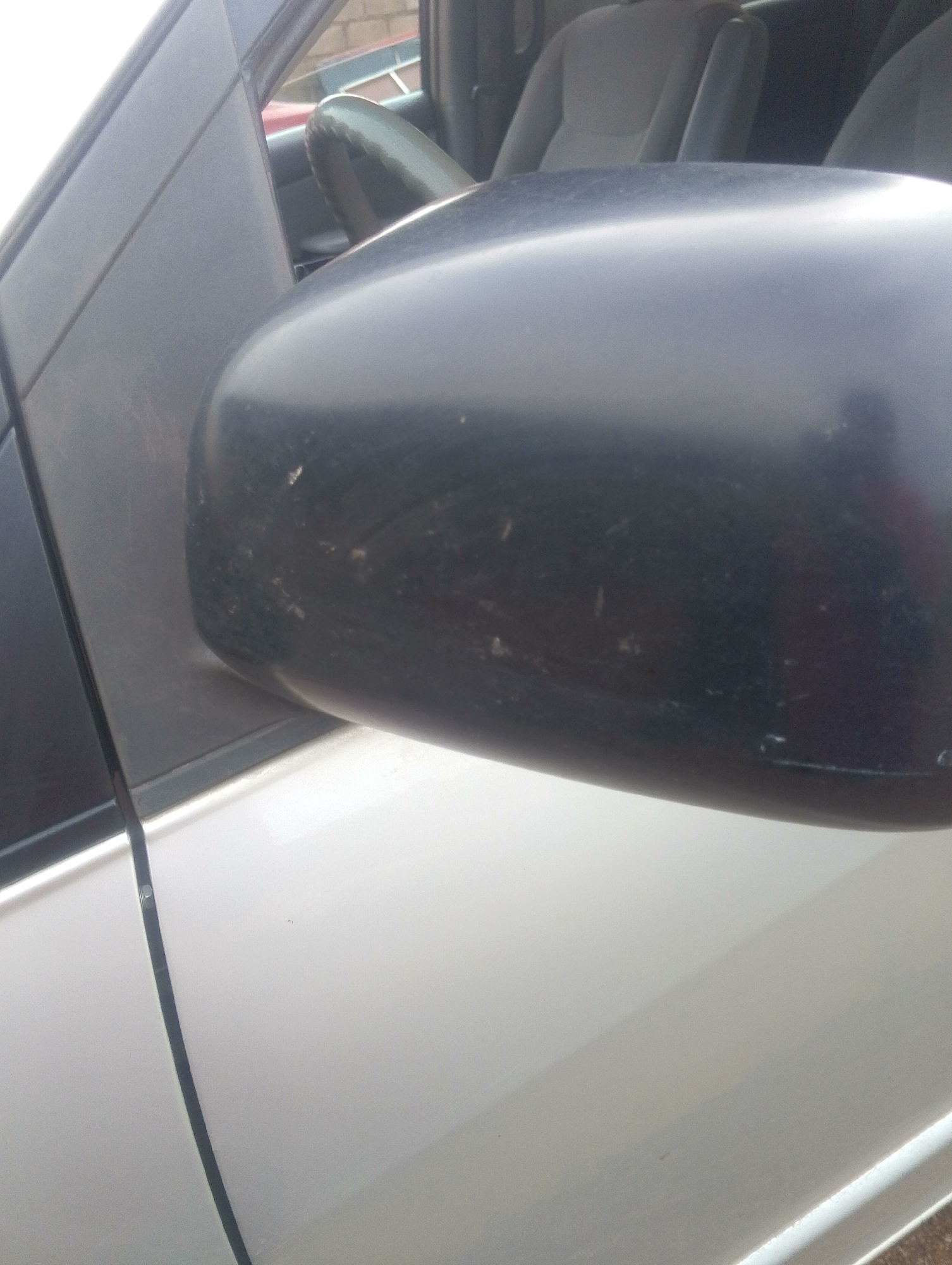 The image displays a close-up view of the side mirror of a silver vehicle. The mirror housing is black and appears to have scuffs and dirt marks. The car's interior, including the seats and a portion of the steering wheel, is visible through the window. The environment suggests an outdoor setting, possibly a street or parking area. The image is a photograph taken in the city of Jos, Nigeria.