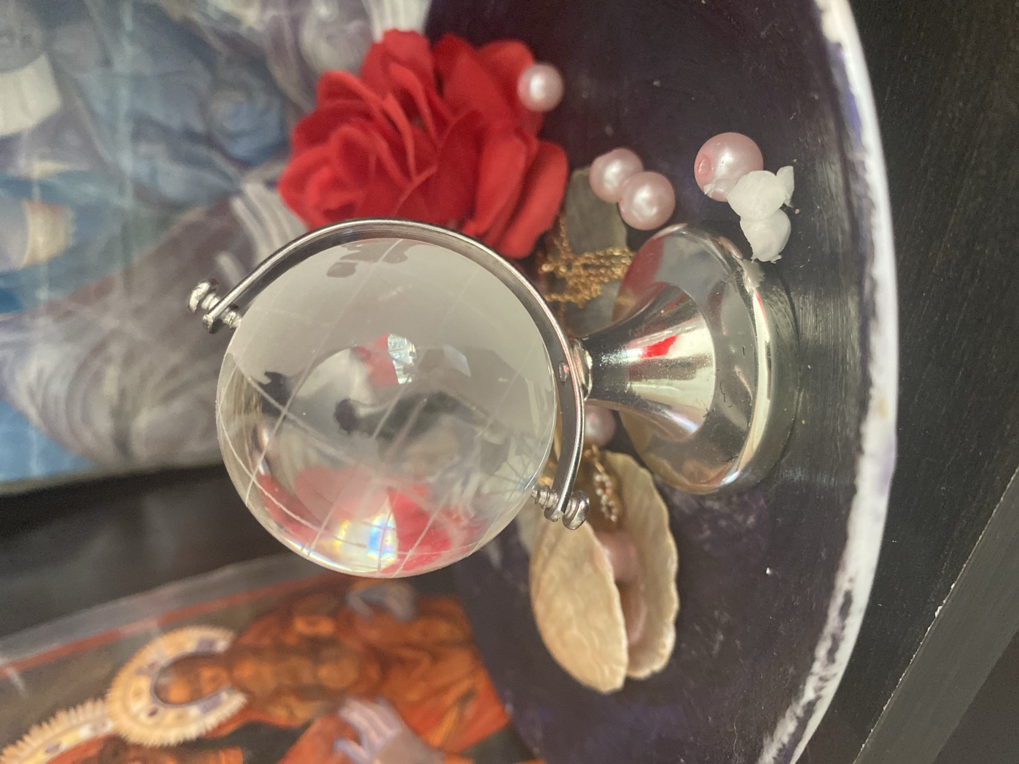 A small, glass globe rests on a silver stand, positioned on a dark, circular tray. The globe reflects light, showing hints of red and blue. Around the globe are several pale pink pearls, a faux red rose, a seashell with a pearl inside, and a delicate gold chain. In the background, a framed image of a person with a halo is visible, along with a blurred background of blue and white. The scene is illuminated by artificial light, casting shadows on the objects. The photograph does not provide any identifiable context related to Blagoevgrad, Bulgaria.