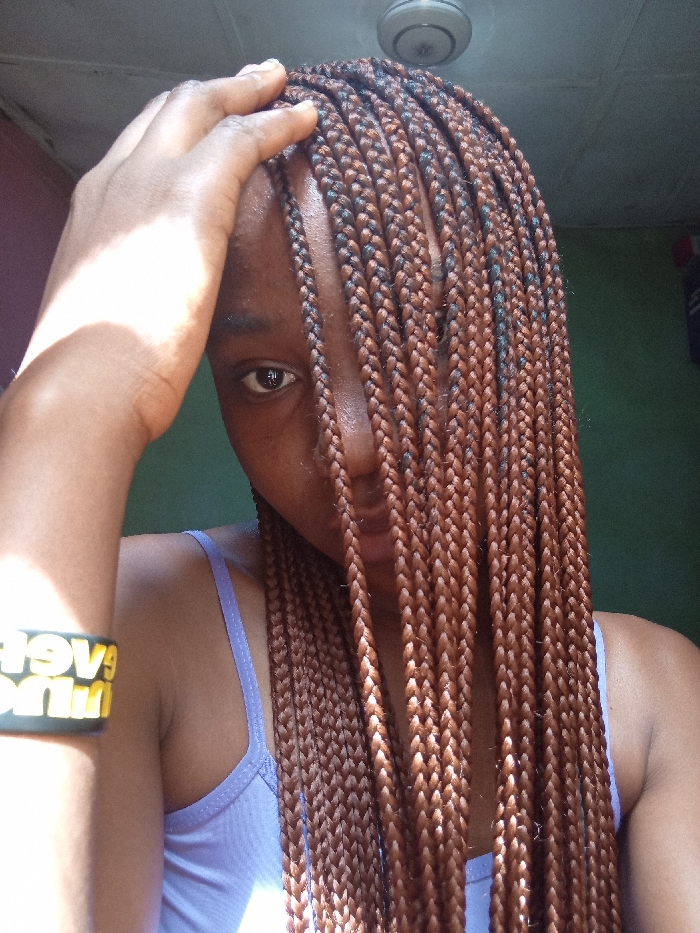 An indoor portrait captures an individual in the city of Gwagwarwa, Nigeria. The subject, an individual with dark skin, is positioned from the chest upwards. Long, small-to-medium sized box braids, predominantly reddish-brown with interwoven darker strands, cascade across the right side of the face, obscuring the right eye, nose, and mouth. The subject's left eye, dark brown, is visible and directed towards the viewer. A dark-skinned left hand rests on the upper forehead/hairline. The subject wears a light lavender spaghetti-strap top. A black wristband featuring inverted yellow text is worn on the left wrist. The background consists of a green-painted wall and a white ceiling with a centrally mounted ceiling fan. Strong, directional light illuminates the left side of the subject's face, hair, and arm, casting the right side into shadow.