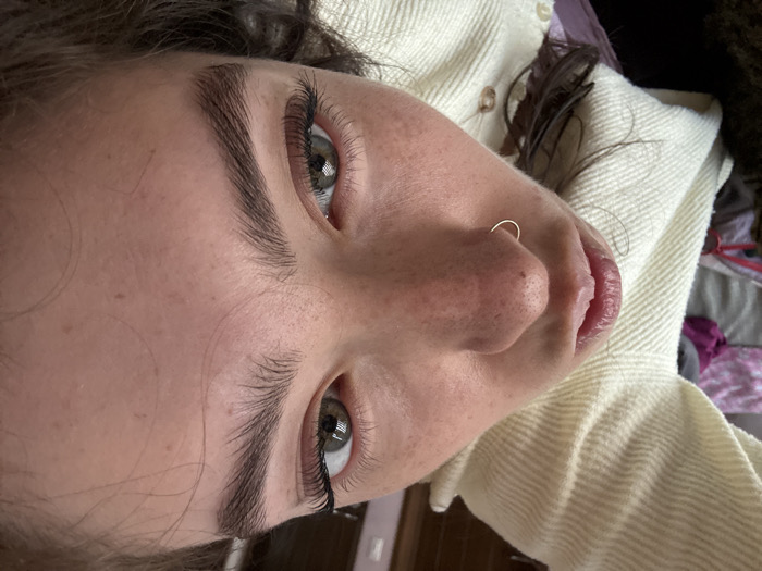 A close-up, rotated perspective of a person's face reveals clear facial features. Dark eyebrows frame green eyes with visible lashes. The person's nose is adorned with a gold hoop piercing. Soft pink lips are slightly parted. The skin exhibits freckles. The individual wears a textured, cream-colored top, with glimpses of what appears to be hair and bedding in the background.
