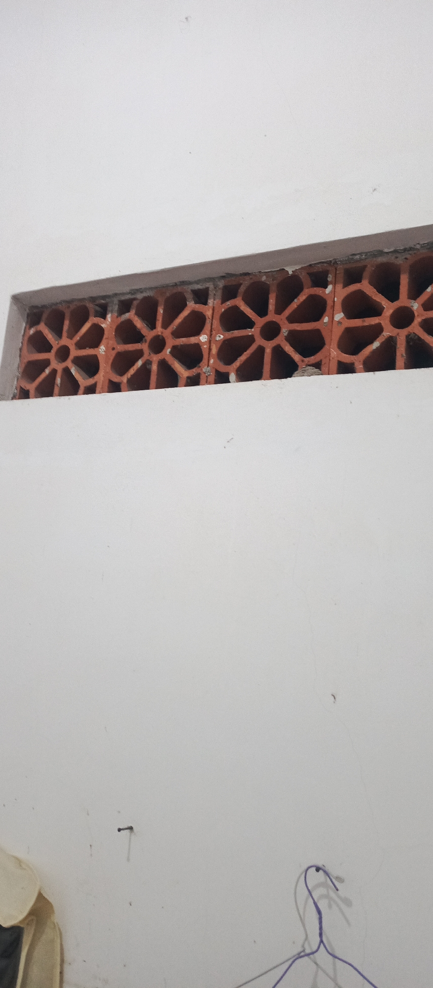 The image shows a close-up of a white wall with a decorative terracotta vent. The vent is rectangular and features a repeating geometric pattern with circular openings. The wall appears to be indoors or on a covered exterior, as there is no visible outdoor environment. The lighting suggests an indoor setting. Below the vent, the wall is bare, with a visible nail or hook on the left side. Towards the bottom right, a partial view of a purple wire hanger is visible, suggesting it is hanging on something out of frame. The overall impression is of a simple, undecorated interior wall space. The location in Arjasa, Indonesia, is noted, but this is not visually evident in the image itself.