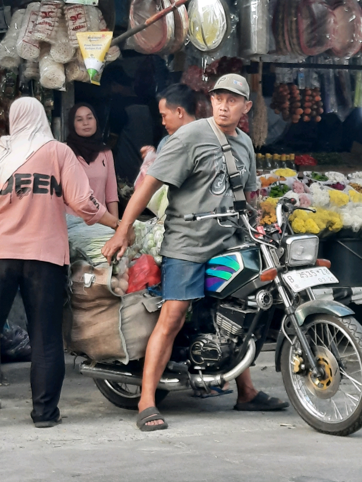 The image depicts a street scene in East Jakarta, Indonesia. A man wearing a grey t-shirt, blue shorts, and a baseball cap is sitting on a green and black motorcycle. He has a bag slung across his chest and is looking to his right. To his left, two women are partially visible. One woman, wearing a pink long-sleeved shirt and black pants, has her back to the camera. The other woman, wearing a dark hijab and a pink top, is looking towards the motorcycle. The motorcycle is laden with large burlap sacks and bags of produce, including what appear to be potatoes and leafy green vegetables. In the background, market stalls are visible with various goods hanging, including what look like crackers, dried food items, and possibly flowers. The ground is paved with concrete. The overall scene suggests a busy market area.