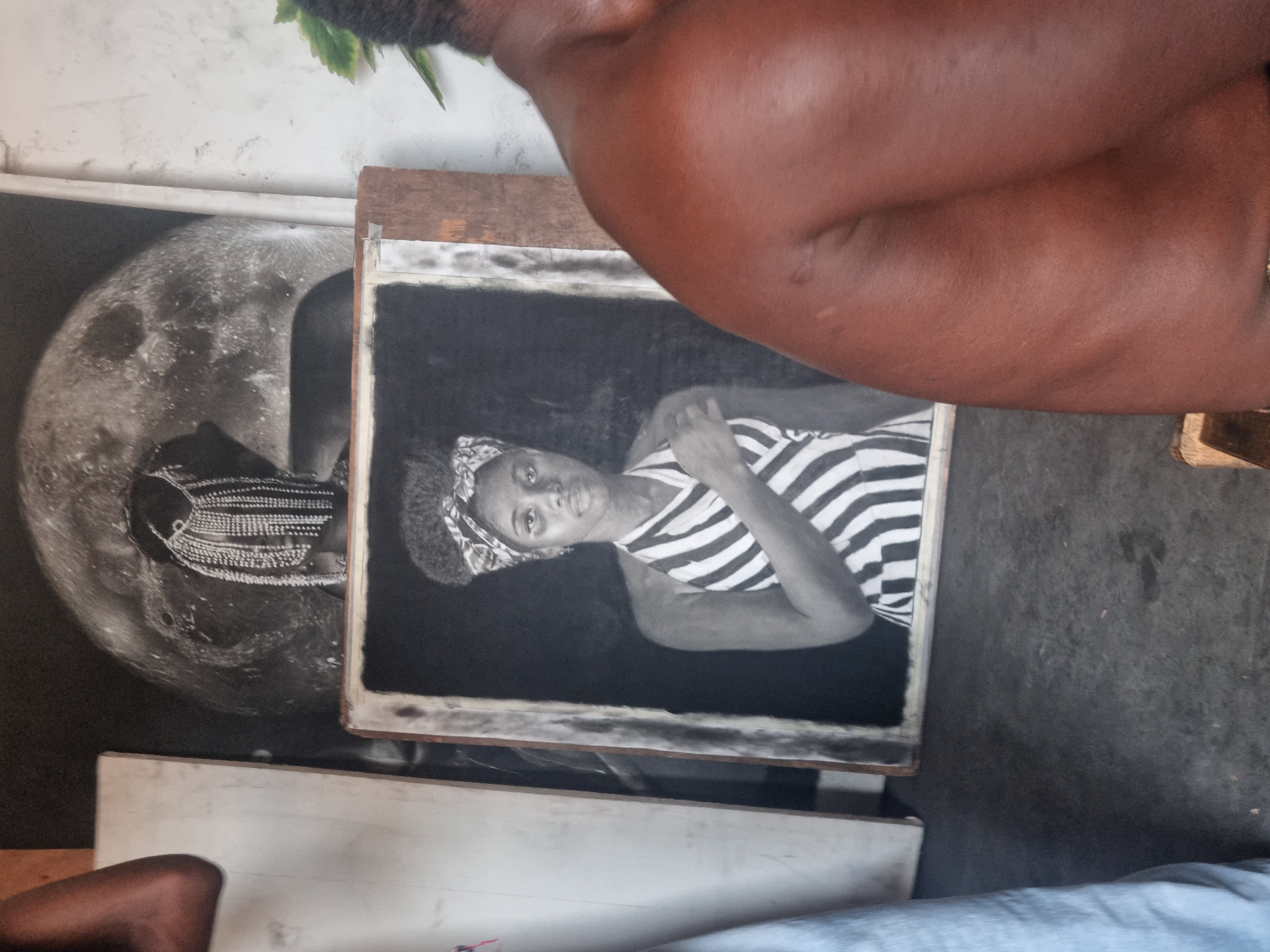 The image displays an artistic scene, likely an outdoor art display or studio in Onitsha, Nigeria. In the foreground, the muscular shoulder and upper arm of a person with dark skin are visible on the right, suggesting they are either an artist at work, a patron, or present in the space.
The central focus is on two prominent artworks, both appearing to be black and white drawings or prints. To the right, a framed portrait depicts a young woman in grayscale, looking directly forward. She wears a patterned headwrap and a striped top. This framed artwork rests on what appears to be a wooden easel or stand.
To the left and slightly behind the framed portrait, a larger, unframed artwork is visible. This piece features a detailed crescent moon, rendered with craters and texture. Partially obscured by the framed portrait, a figure wearing elaborate, intricately patterned traditional attire is visible beneath the moon, suggesting a connection to cultural themes.
The background shows a plain, light-colored wall that appears somewhat weathered, and the ground is a dark surface, possibly concrete or dirt. The lighting suggests it is daytime. There is faint, illegible text on the wooden frame of the portrait and potentially on the moon artwork, but it is not clear enough to decipher. The overall impression is one of a creative space, possibly an informal gallery or an artist's workspace.