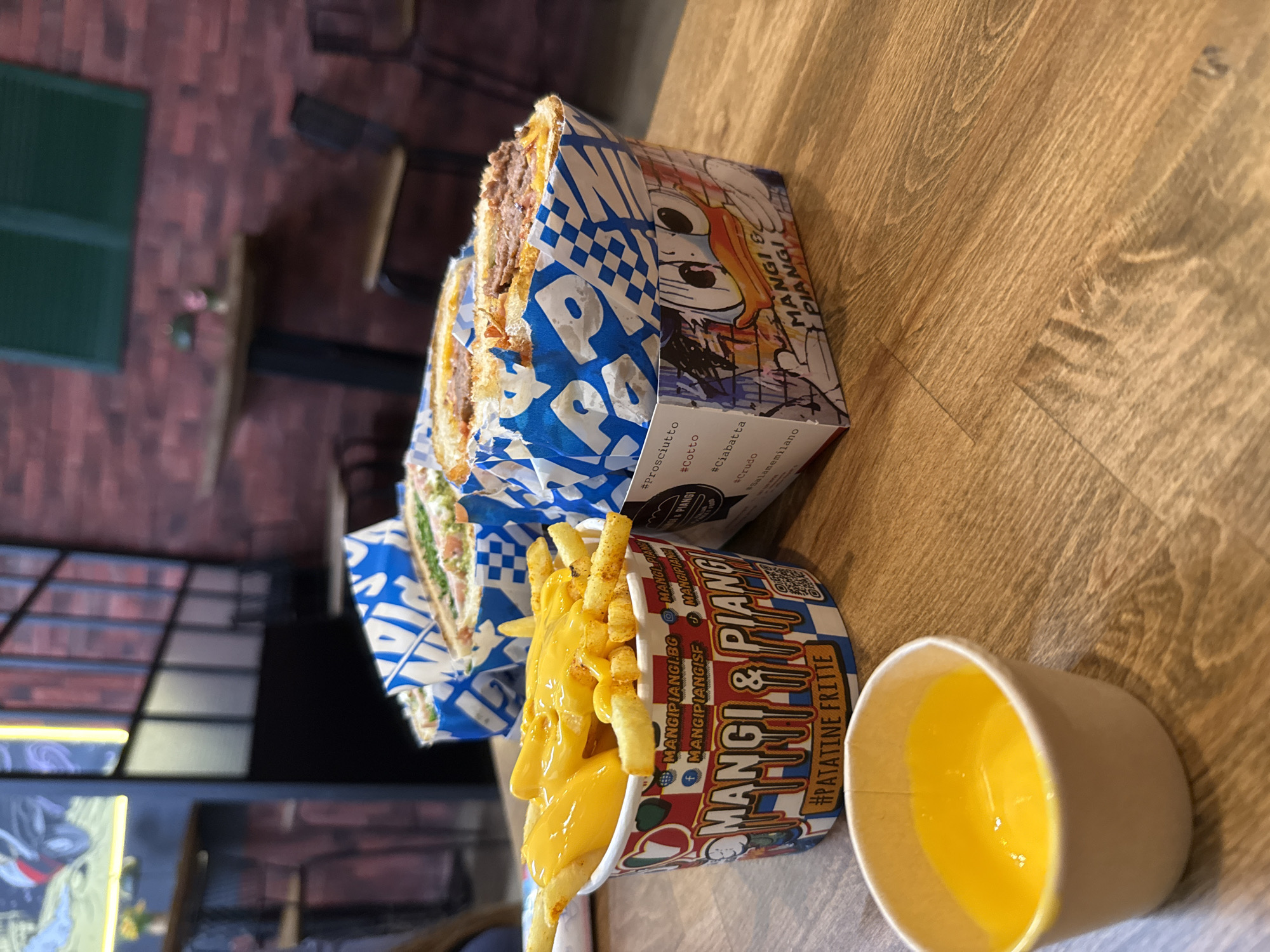 The image shows a meal from the restaurant "Mangi & Piangi", presumably located in Sofia, Bulgaria, based on the text “MANGIPIANGI.BG” visible on the French fries container. A sandwich, packaged in blue and white checkered paper, is situated next to a container of French fries covered in cheese sauce. A small cup of yellow sauce is positioned to the right of the fries. The meal is placed on a wooden surface.