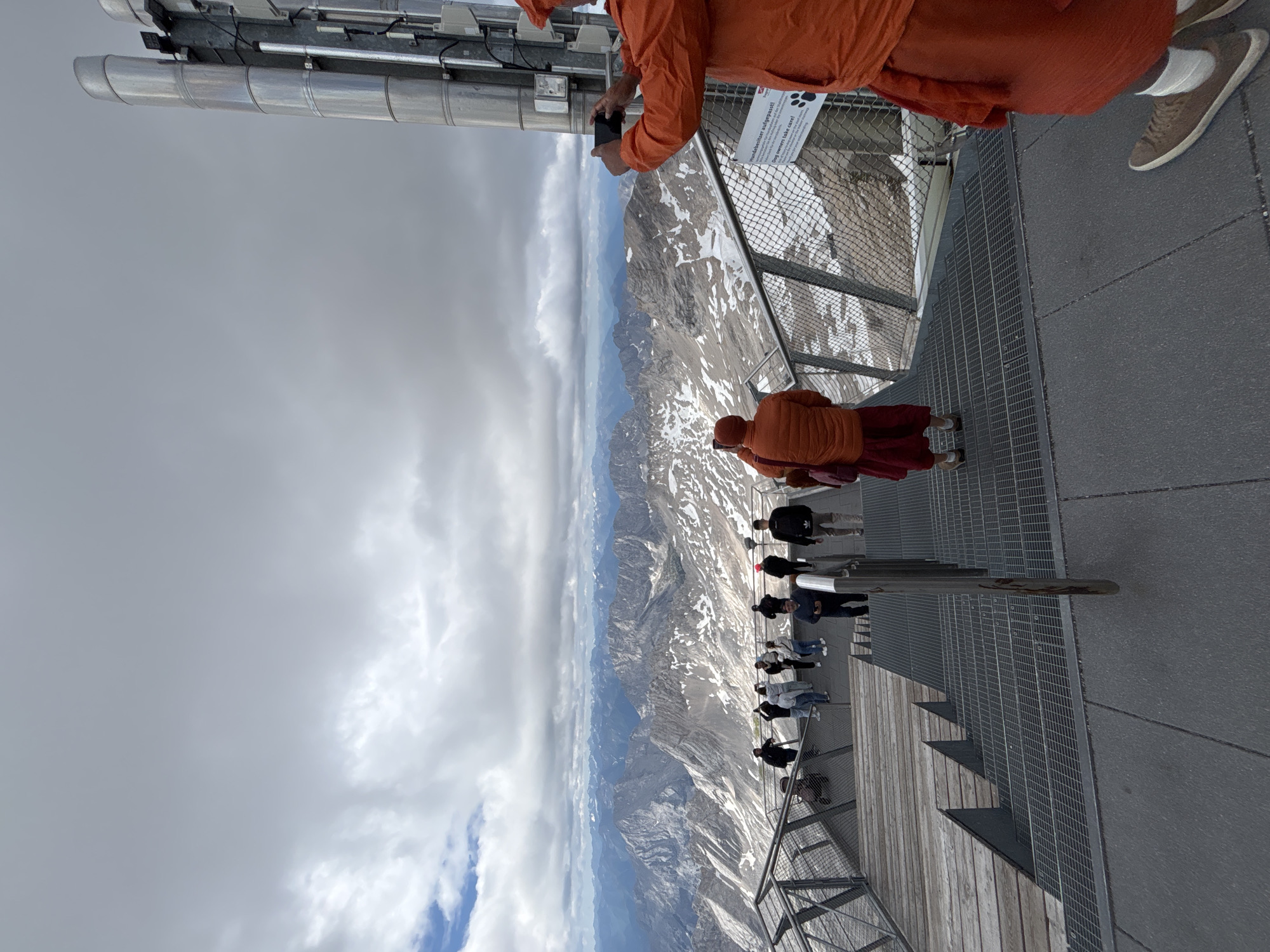 The image depicts a view from an elevated platform overlooking a vast mountain range, likely the Alps, with snow-capped peaks under a cloudy sky. The platform itself is constructed with metal grating and wooden planks, featuring railings and metal stairs leading downwards. Several people are visible on the platform, some looking at the scenery, others walking down the stairs, and one individual in an orange robe is taking a photograph with a smartphone. Another person in a similar orange robe is in the foreground, also holding a phone. There is a sign on a pole in the background with German text and an image of paw prints, indicating a rule related to dogs. The overall scene suggests a tourist viewpoint in a mountainous region. While the exact location is not explicitly stated, the context of Garmisch-Partenkirchen, Germany, known for its proximity to the Zugspitze, Germany's highest peak, makes it a plausible setting.