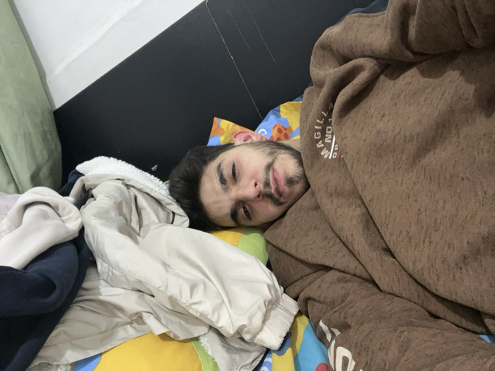 A man lies in bed surrounded by clothes. His face is visible, with eyes partially closed. He is partially covered by a brown hooded sweatshirt. A pile of clothing, including a tan jacket with a white lining and a dark blue garment, sits beside him. The bed has a yellow and blue patterned sheet. The headboard is black. A light green curtain is visible in the upper left corner. The lighting is dim.