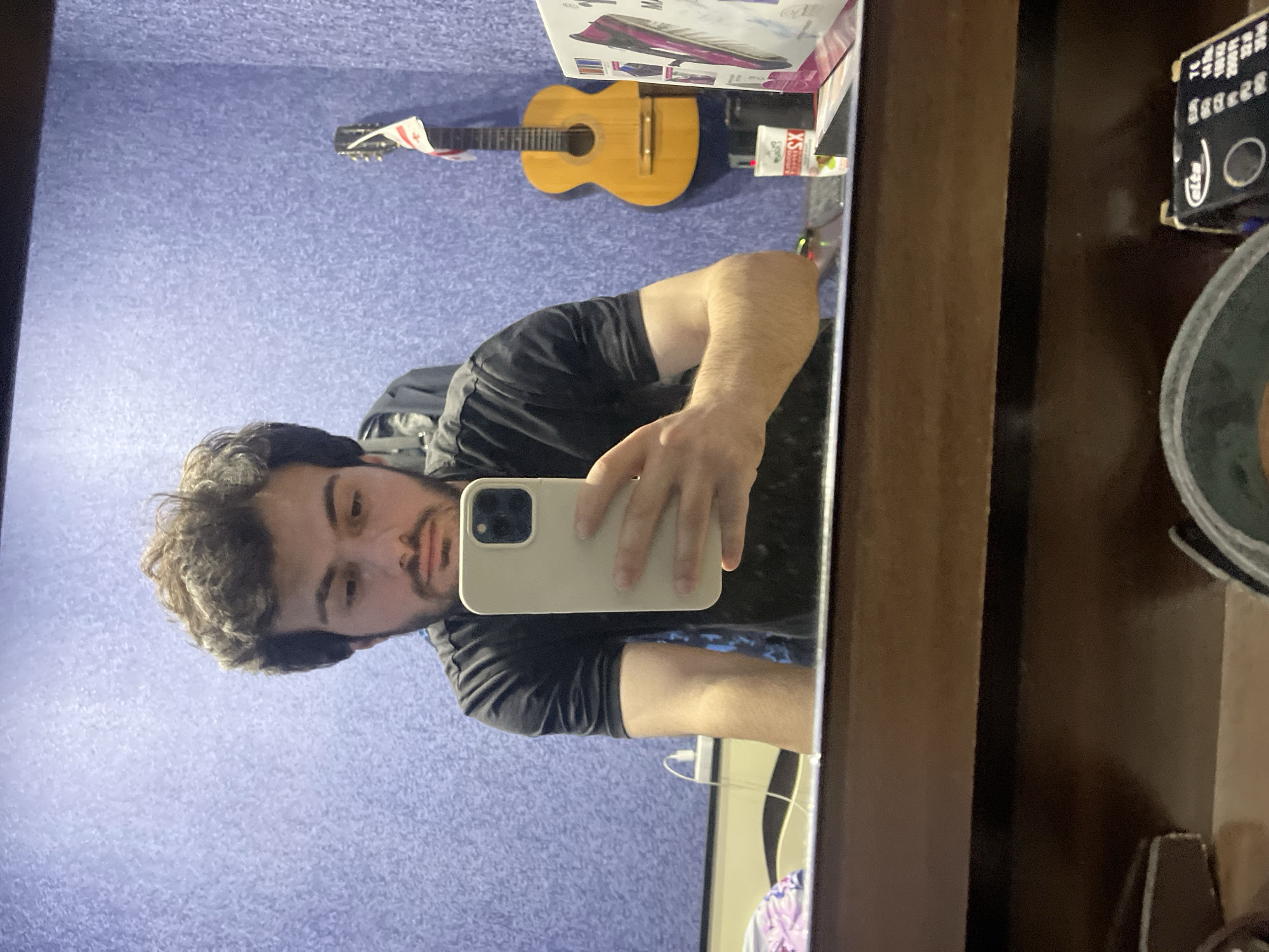 The image, taken in Tbilisi, Georgia, shows a young man with curly dark hair and a beard, wearing a black t-shirt, taking a mirror selfie. He is holding a light-colored smartphone in his right hand. The background features a textured purple wall. To the right of the man, a classical guitar with a Georgian flag attached to its headstock hangs on the wall. On a surface in front of the wall, a white cup with "SX" visible and a box with a pink and purple design are present. The foreground of the image is dominated by the reflection of a wooden surface, likely a desk or table, with various objects visible in the reflection, including what appears to be a spool of wire and a small black box with text. The lighting suggests an indoor setting, possibly a bedroom or studio.