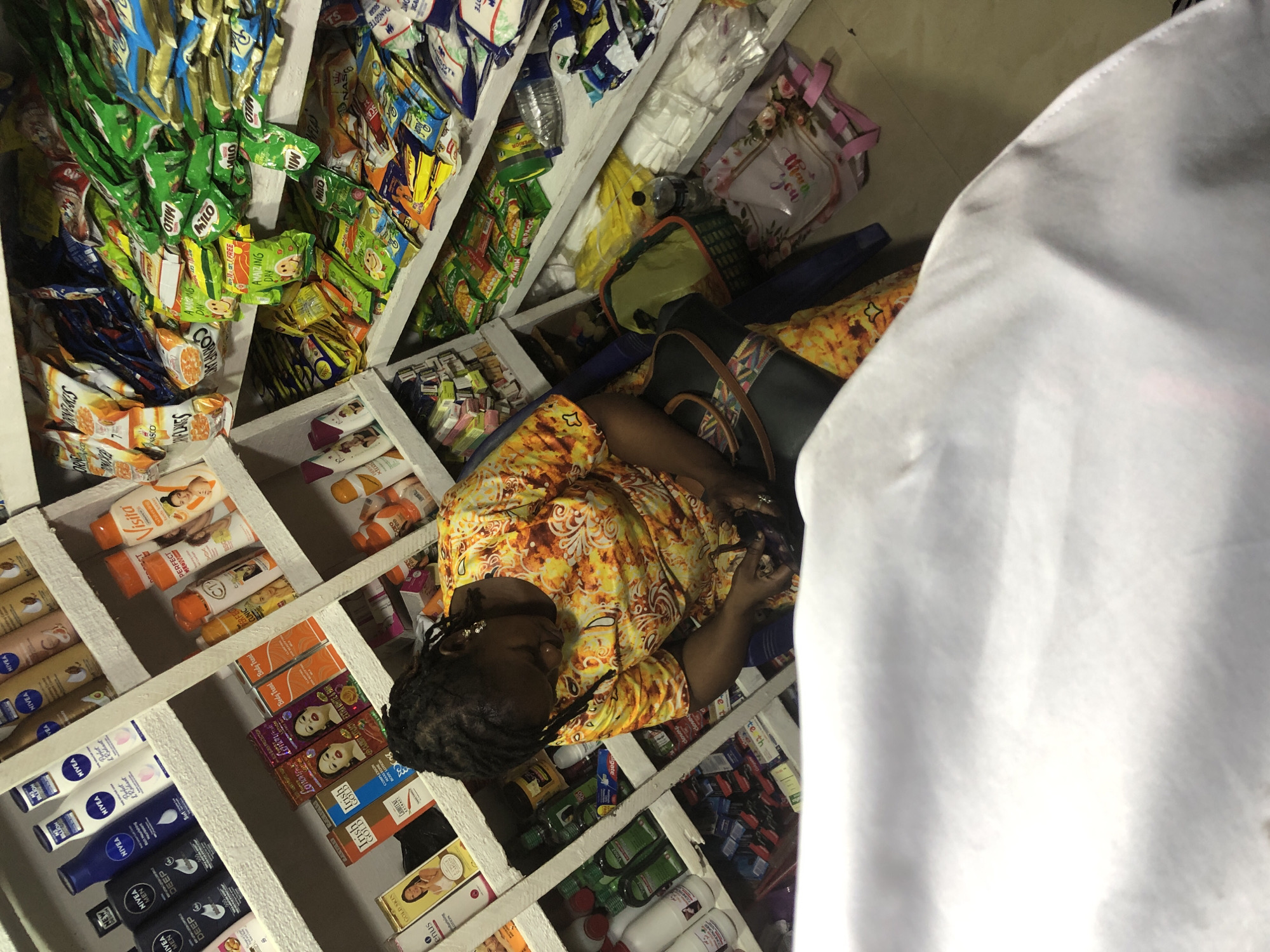 In a crowded stall within Ado, Nigeria, a woman with dark, braided hair is engrossed in examining an item in her hands. She is wearing a vibrant, yellow, orange, and white patterned dress. Her hair is tied back, and she has earrings in her ears. The stall, packed with various goods, features shelves overflowing with an assortment of products.
On the shelves, a diverse range of items is visible. These include toiletries like Nivea lotions, Visita, and other beauty products. Nestled amongst them are food items, such as Milo sachets, Kellogg's corn flakes, and other snacks in colorful packaging. There are also several bottles of liquid, possibly cleaning agents or tonics, and boxes of toothpaste and other personal care items.
The woman is seated on the floor or a low stool, with a black and multi-colored bag resting beside her. The floor is tiled, and various plastic bags and containers are scattered around the stall. The lighting suggests an indoor environment, possibly a small shop or market stall. The scene conveys a sense of commerce and daily life within the bustling city.