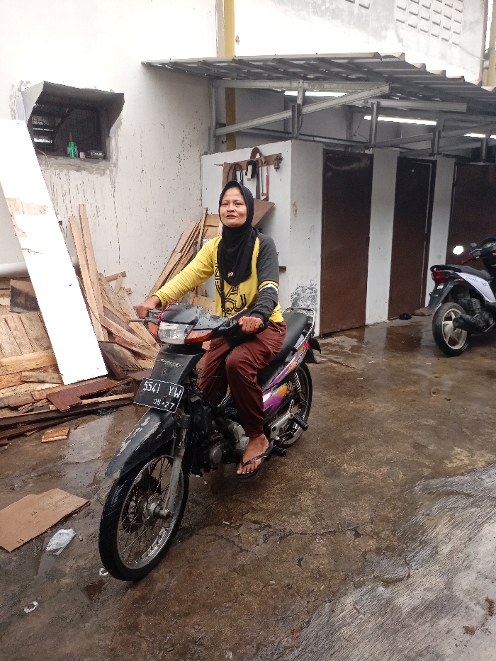The image shows a woman sitting on a black motorcycle in a grey, concrete driveway. She is wearing a yellow long-sleeved shirt, brown pants, and a black headscarf. She is looking at the camera and smiling. The motorcycle has a black license plate that reads "5541 YAW 05-27." The motorcycle is parked in front of a building with a metal roof. There are two wooden doors to the right of the motorcycle. To the left of the motorcycle is a pile of wood scraps. The scene appears to be in Katapang, Indonesia, as indicated by the provided coordinates. The ground is wet and the weather is overcast.