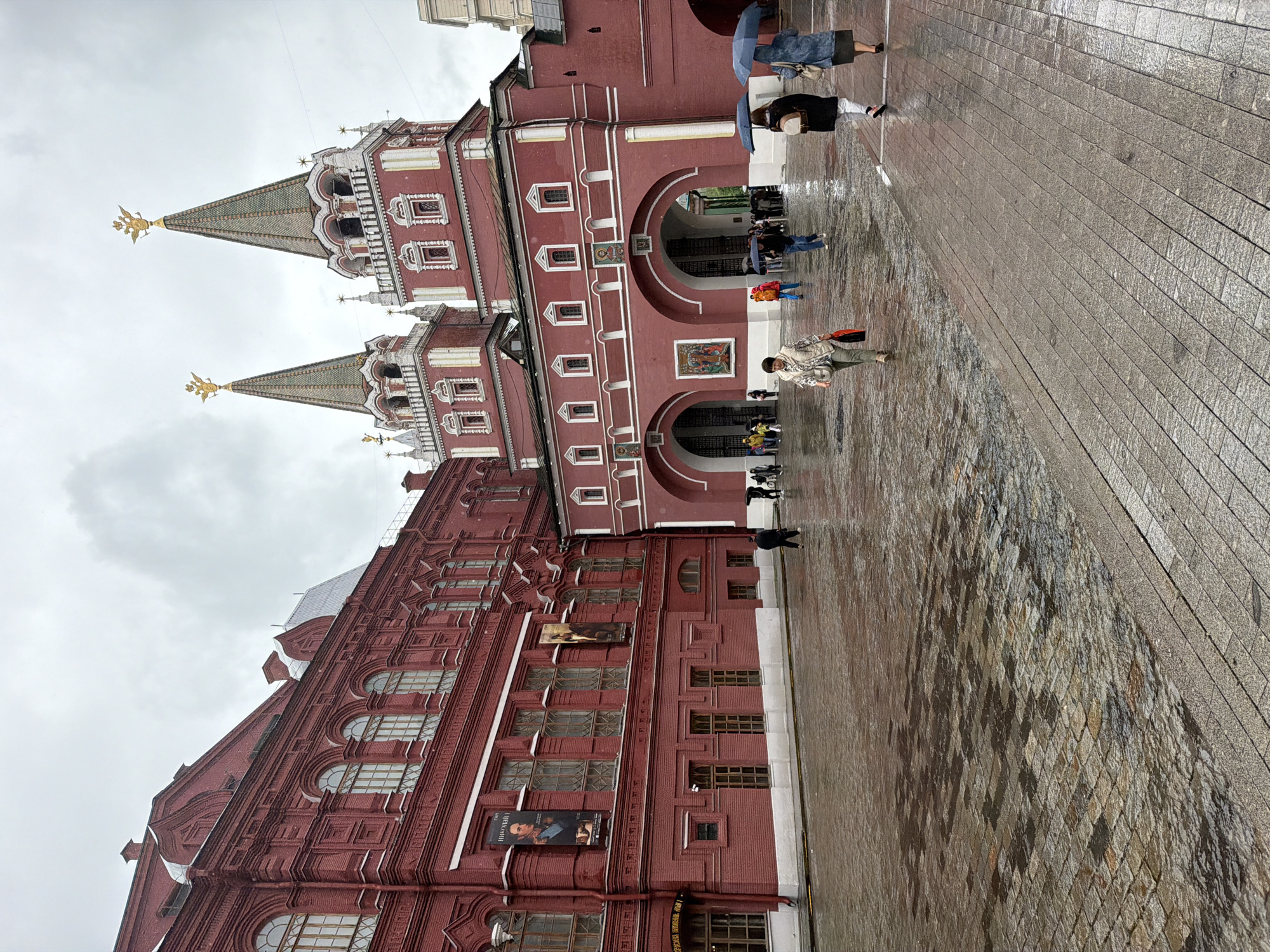 The image depicts a view of the State Historical Museum and the Voskresensky Gate in Moscow, Russia, on a cloudy and rainy day. The red brick facade of the museum dominates the left side of the frame, featuring arched windows and two large banners with portraits. To the right, the Voskresensky Gate, also red brick with decorative white accents and intricate detailing, stands prominently. It has two prominent, pointed towers topped with golden double-headed eagles, symbolizing Russia. The ground in the foreground is paved with wet cobblestones, reflecting the overcast sky. Several people are present, walking under umbrellas. Three women are walking from right to left in the foreground. The woman on the far right holds a blue umbrella and wears a denim jacket over a dark skirt. The woman in the middle has a backpack and is also holding a dark umbrella. The woman on the left walks away from the camera, carrying a red bag and wearing a light-colored jacket and pants. In the middle ground, a man in a bright orange jacket walks towards the gate, and other figures are seen approaching or standing near the arches of the gate. The overall atmosphere is somber due to the weather, but the architecture provides a vibrant visual contrast.