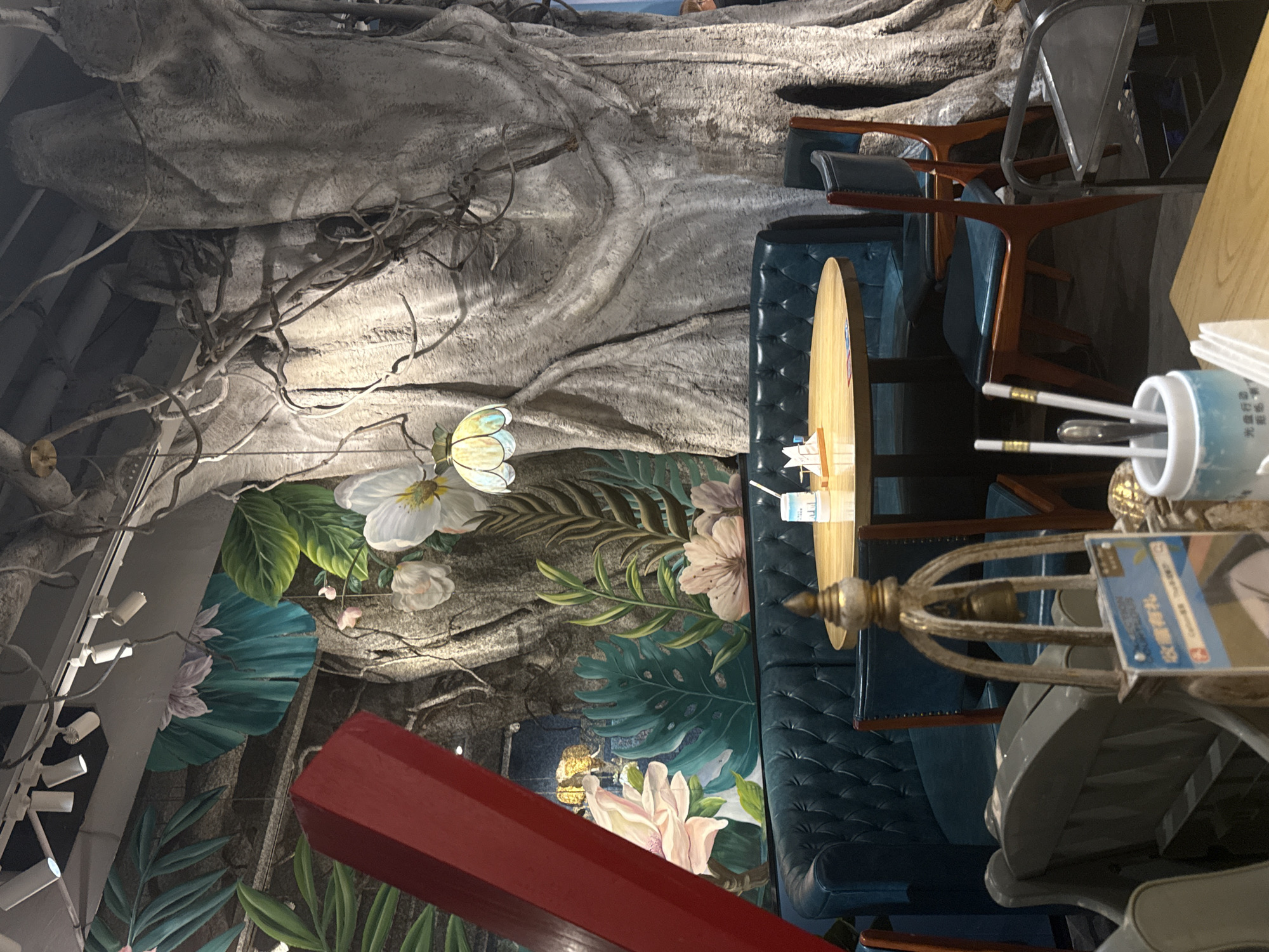 The image depicts the interior of a restaurant in Yiwu, China, with a distinct jungle-themed decor. A large, artificial banyan tree dominates the background, with its gnarled roots and branches integrated into the wall. Lush green and vibrant floral murals adorn the walls, featuring large leaves and blossoms, creating an immersive botanical atmosphere.
In the foreground, a plush, teal-colored tufted booth seating arrangement curves around a circular wooden table. On the table, a white paper dispenser with a small flag, a clear plastic cup with a straw, and what appear to be chopsticks in a holder are visible. Two wooden chairs with teal seats are positioned next to the table.
A decorative element, possibly a lamp or sculpture, with a golden base and a bell hanging from an arched frame, stands in front of the booth. A red, rectangular structural element is partially visible in the upper left quadrant, leaning into the frame. The lighting in the restaurant appears to be a mix of spotlights on the ceiling and a decorative pendant lamp with a stained-glass-like shade hanging from the artificial tree. The overall impression is of an exotic and atmospheric dining space.