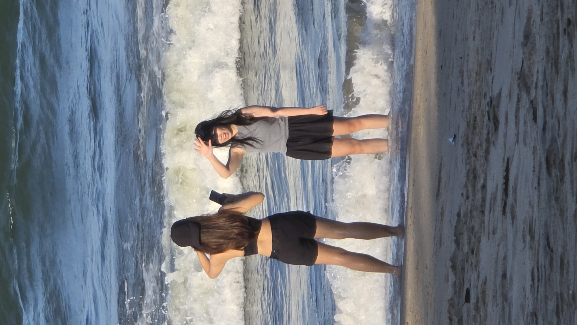 The image depicts two young women standing in shallow ocean water on a sandy beach. The location appears to be Nha Trang Ward, Vietnam, based on visual characteristics of the water and beach. The top woman waves, while the bottom woman is taking a photo with a black camera. The water is choppy with small waves, and the sand is wet and textured. A small white object and a dark object are also visible on the sand.