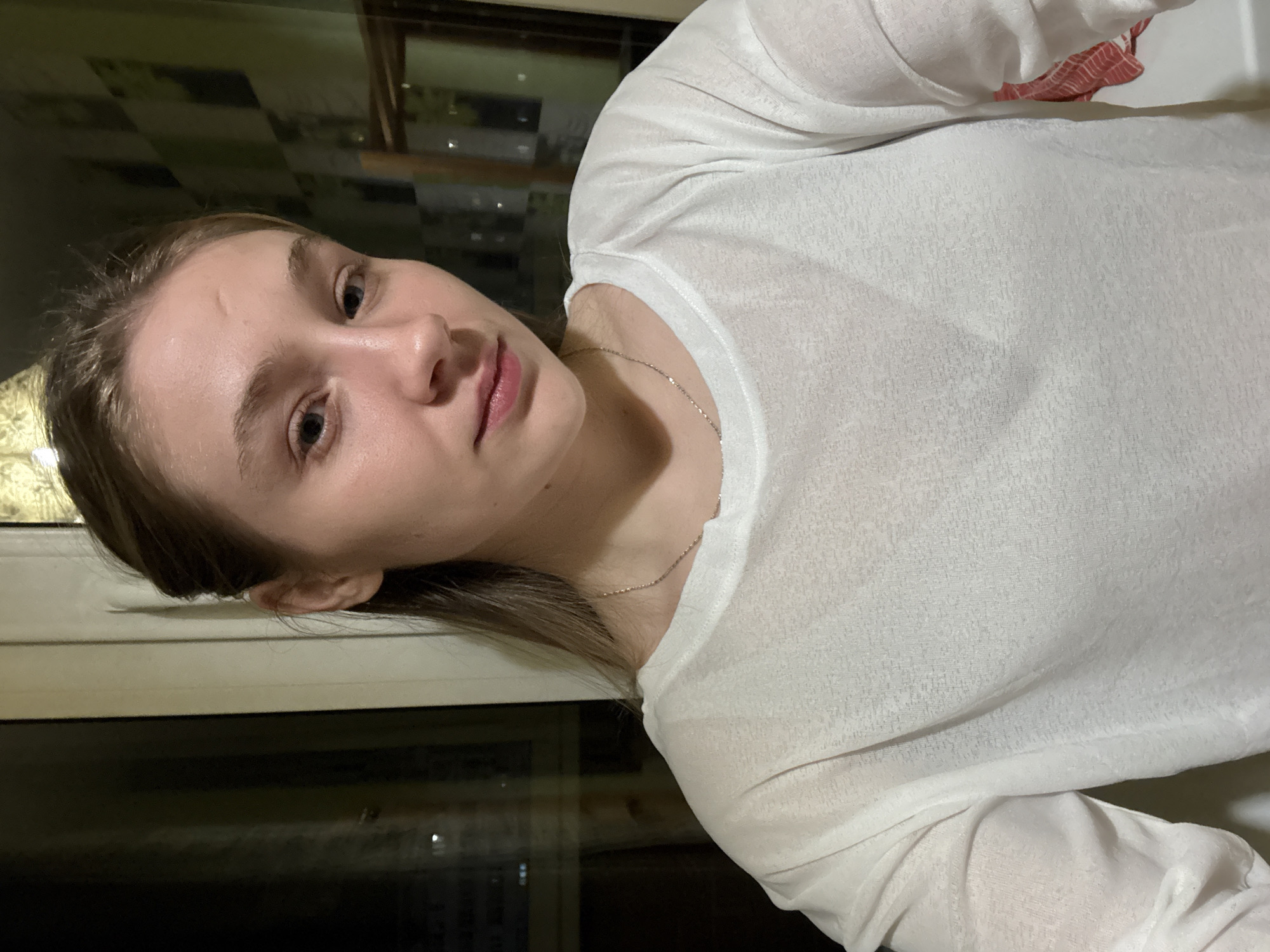 A woman is indoors, near a window, wearing a white long-sleeved shirt and a thin silver necklace. Outside the window is a cityscape at night. While I cannot specifically confirm that the location is in Izhevsk, Russia without further information, the scene shows a person indoors with a window view of a city at night.
