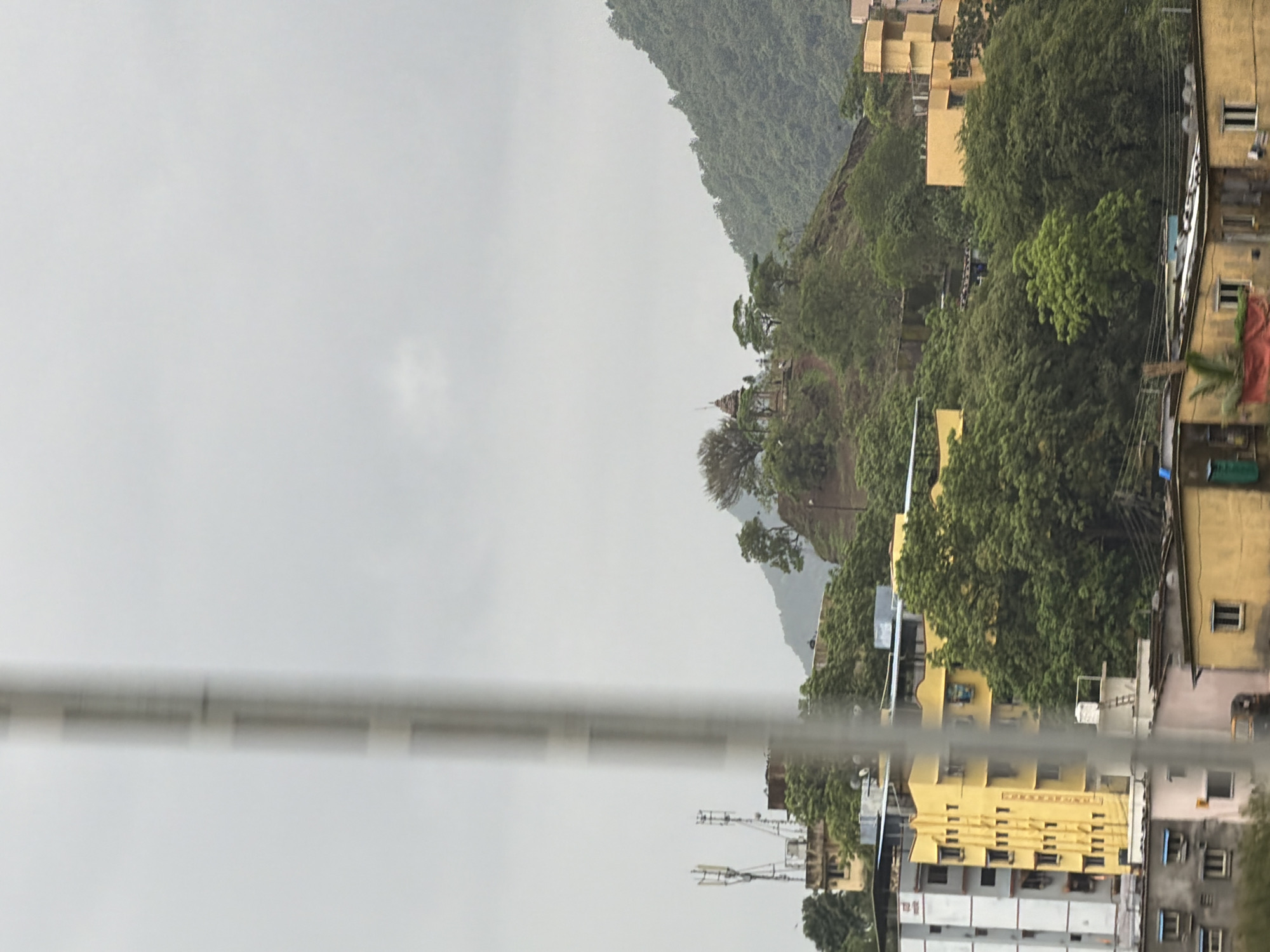 The image depicts a cloudy day in Vasai-Virar, India, with a focus on a residential area nestled against a lush green hillside. In the foreground, several buildings are visible, including a tall, white and gray apartment building with numerous windows and balconies, and a vibrant yellow, multi-story structure with a distinctive architectural design. Adjacent to these are lower-lying, older buildings with various window styles and some visible utilities like satellite dishes.
Further up the hill, more buildings are scattered amongst the dense foliage. A prominent yellow building with a unique, stepped facade stands out. The hillside itself is covered in a thick carpet of trees, with some darker, more rugged areas suggesting steeper inclines or rock formations.
At the crest of a prominent hill, a small, weathered temple or shrine structure is visible, topped with a spire. The surrounding trees on this elevated area are sparser, allowing the structure to be seen more clearly. Two tall communication towers, likely for cellular service, are located on a lower ledge of the hill, to the left of the temple.
The sky is overcast with a uniform layer of gray clouds, obscuring any direct sunlight and casting a diffused light over the entire scene. A thin, vertical, out-of-focus pole-like object runs through the center of the image, partially obscuring the view of the buildings and landscape behind it. The overall impression is one of a densely populated urban environment encroaching on a natural, hilly landscape under a somber sky.