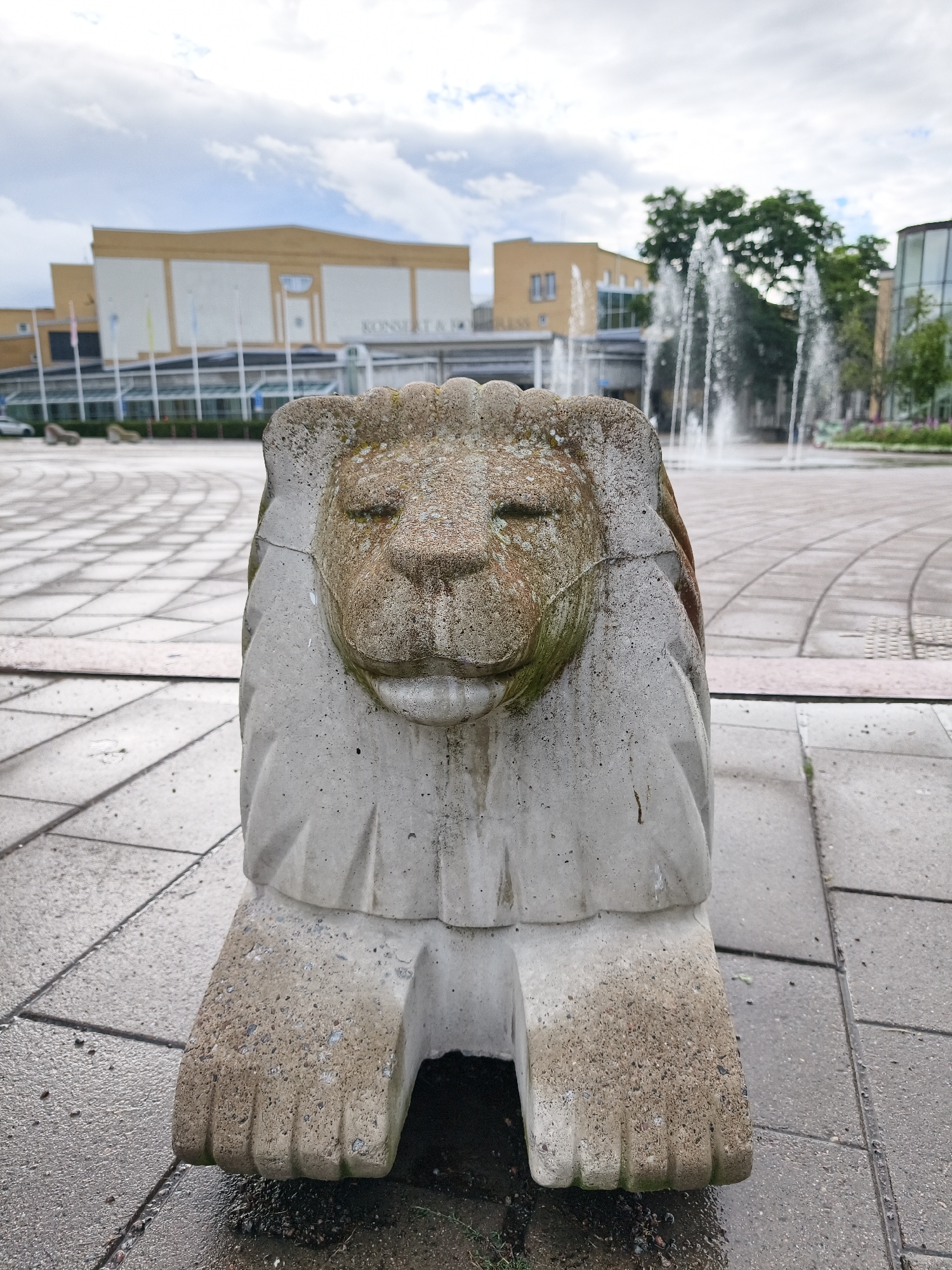 The image shows a concrete sculpture of a lion in the foreground, located in a paved public square in Linköping, Sweden. The lion sculpture is weathered and has some moss growth on it. In the background, there is a large, modern building with the text "KONSERT &..." visible on its facade, suggesting it is a concert hall. A fountain with several streams of water is active in the middle ground. The square is paved with square tiles arranged in a circular pattern, and the ground is wet, indicating recent rain. The sky is cloudy. The overall environment appears to be an urban public space.