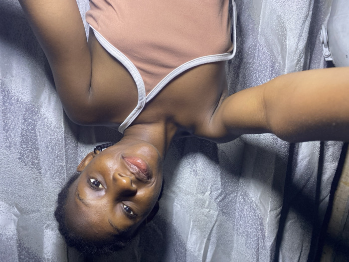 The image shows a single young female, likely an adult, located indoors in Iranla, Nigeria. She has dark skin and her hair is styled in braids. She is wearing a light brown or peach-colored sleeveless top with white trim along the neckline and straps. A black hoop earring is visible in her left ear. The individual is positioned facing the camera, with her head slightly tilted to her left, and she displays a subtle smile. Her right arm is visible in the lower left portion of the frame, extended, suggesting she is holding the device used to capture the image. The background consists of a light gray or silverish textured material, possibly fabric or a patterned wall, with visible shadows indicating an overhead or directional light source.