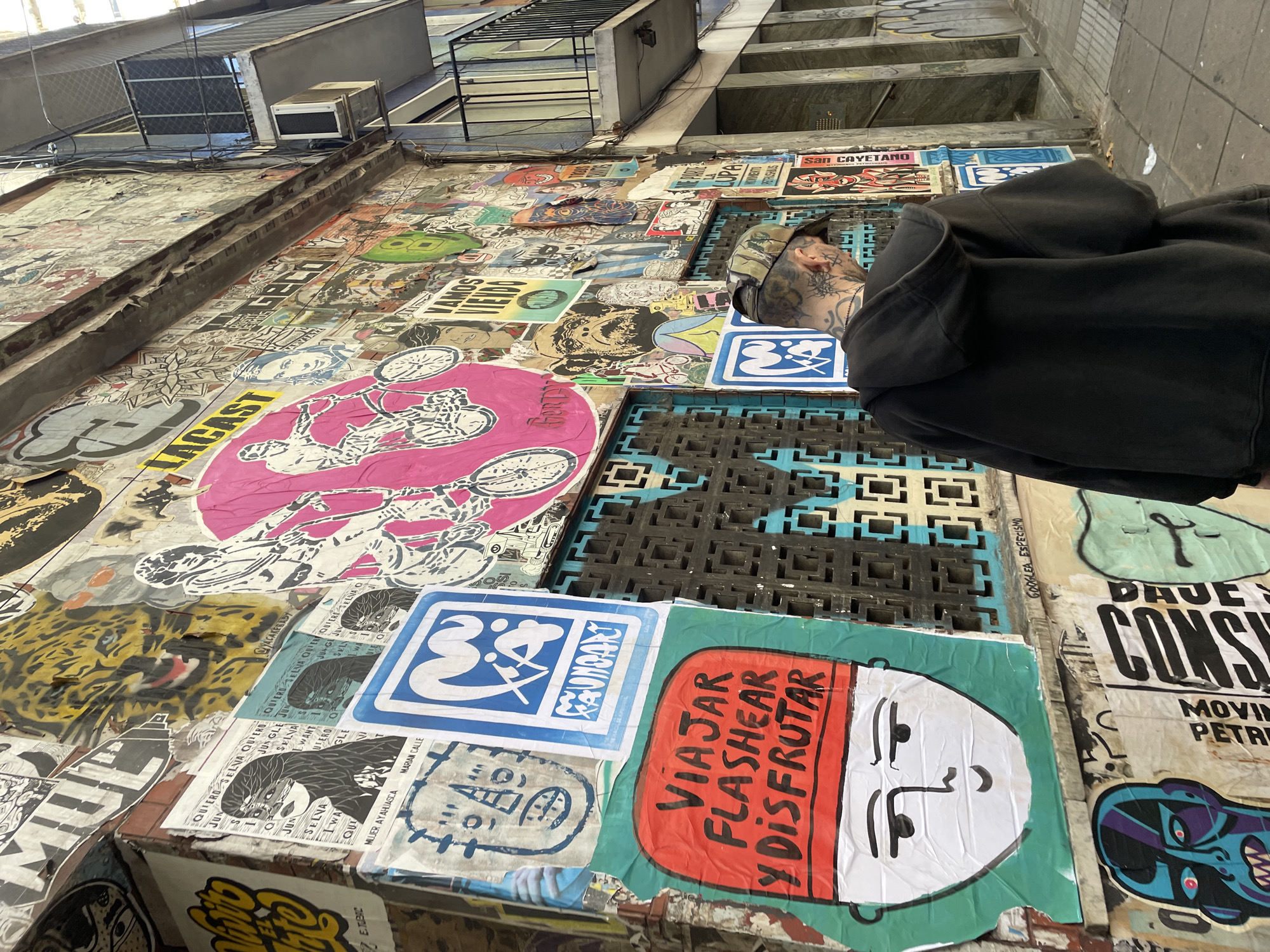The image presents an urban scene in Buenos Aires, Argentina, captured from a low, tilted angle looking upwards. The primary subject is a person, partially visible on the right, wearing a black hoodie and a cap. Notable tattoos adorn their neck, face, and a visible hand, suggesting a distinct personal style.
The setting is an alley or a narrow outdoor space, characterized by a wall or ground surface densely covered with a vibrant collage of street art, stickers, and posters. These artworks vary widely in style and color, creating a dynamic visual tapestry. Among the identifiable elements are a large pink sticker depicting a figure on a bicycle with the text "LACAST," a blue and white sticker featuring a stylized figure and the word "GAUNDART," and a red and white poster with a face and the Spanish phrase "VIAJAR FLASHEAR Y DISFRUTAR" (Travel, Flash, and Enjoy). Other discernible text fragments include "SAN CAYETANO," "DAUE CONS," "QUIERO SELVA," "JUNGLE JUN," "MOVIN PETR," "MUJER AYAHJASCA," "MARIAN CALLE," and "GOOGLEA ESPECISMO." A leopard print stencil and various abstract graphics also contribute to the layered street art.
In the background, multi-story buildings with concrete facades, metal grates, and an air conditioning unit are visible, suggesting a typical urban environment. Exposed wiring runs along the upper structures. The scene is brightly lit, indicating daytime. The activity depicted is primarily the presence of intricate street art and the casual stance of the person, whose identity or full activity is not explicitly clear.