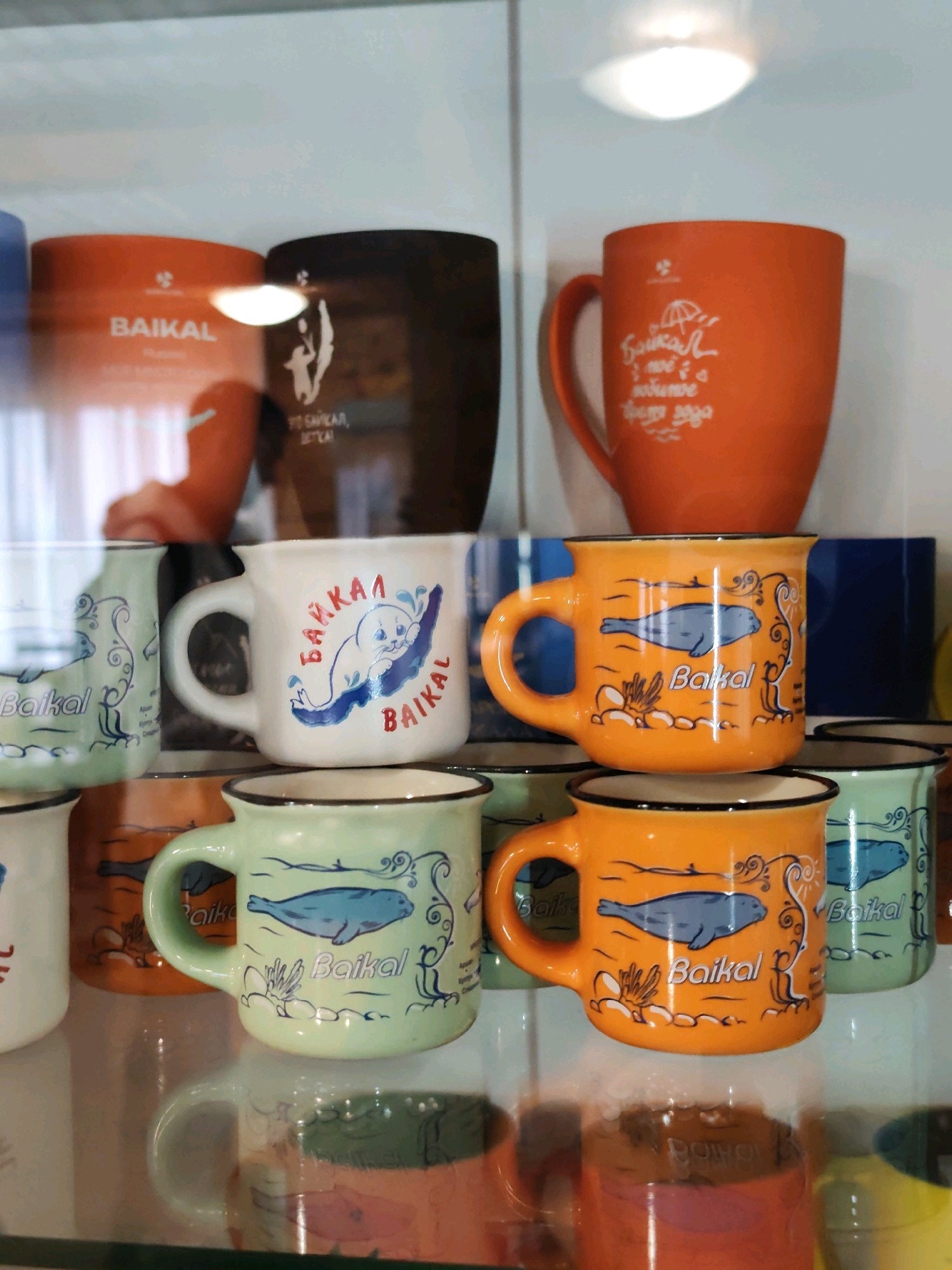 The image shows a display case filled with ceramic mugs featuring designs related to Baikal, a large freshwater lake in Siberia, Russia. The mugs are in various colors, including orange, light green, and white, and some have black rims. Several mugs depict seals and aquatic plants, with the word "Baikal" or "Байкал" written on them in Cyrillic or Latin script. In the background, larger mugs or containers with "Baikal" branding are visible, along with what appears to be a depiction of a person in silhouette. The lighting suggests an indoor environment, possibly a souvenir shop or a museum display. There are no people in the immediate foreground or visible in detail. The specific location of Nikola, Russia, is not directly identifiable from the visual elements within the image itself, as no geographical markers are present.