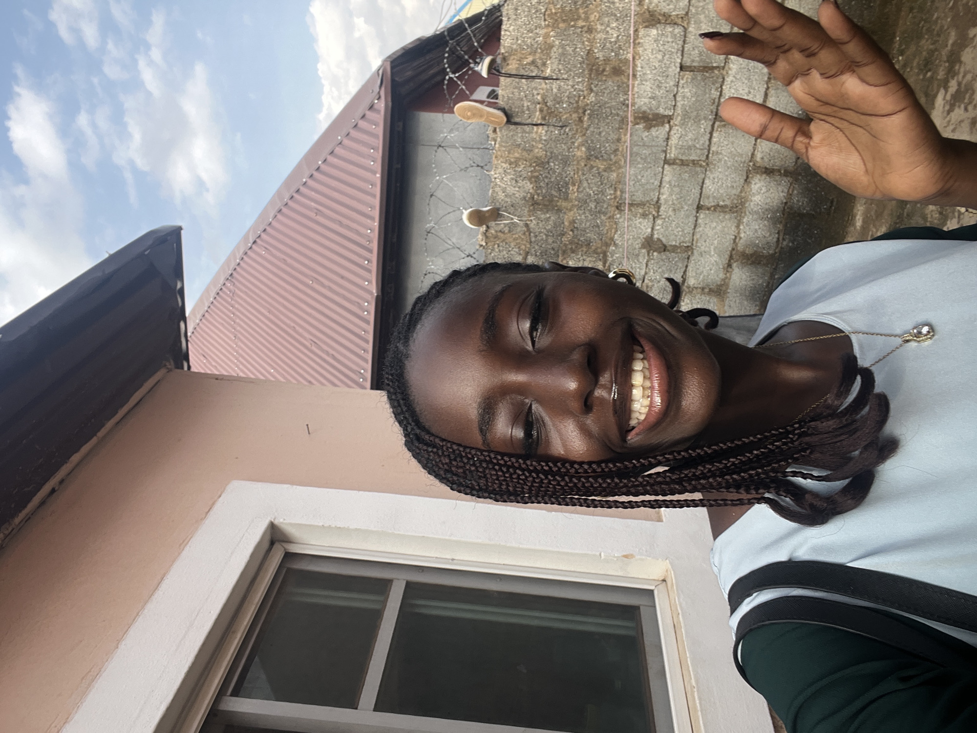 The image is a selfie taken outdoors, likely in a residential area of Abuja, Nigeria. The background consists of a light pink building with a white-trimmed window on the left, and a corrugated metal roof of a building in the middle ground. A grey brick wall runs across the background, topped with barbed wire. A pair of old sneakers are tied to the barbed wire.
The subject is a young Black woman with braided hair, wearing a light blue shirt over a dark green garment. She is smiling and looking directly at the camera, with her right hand raised. She is wearing a necklace with a pendant and hoop earrings. The lighting suggests it is daytime with the sun out.