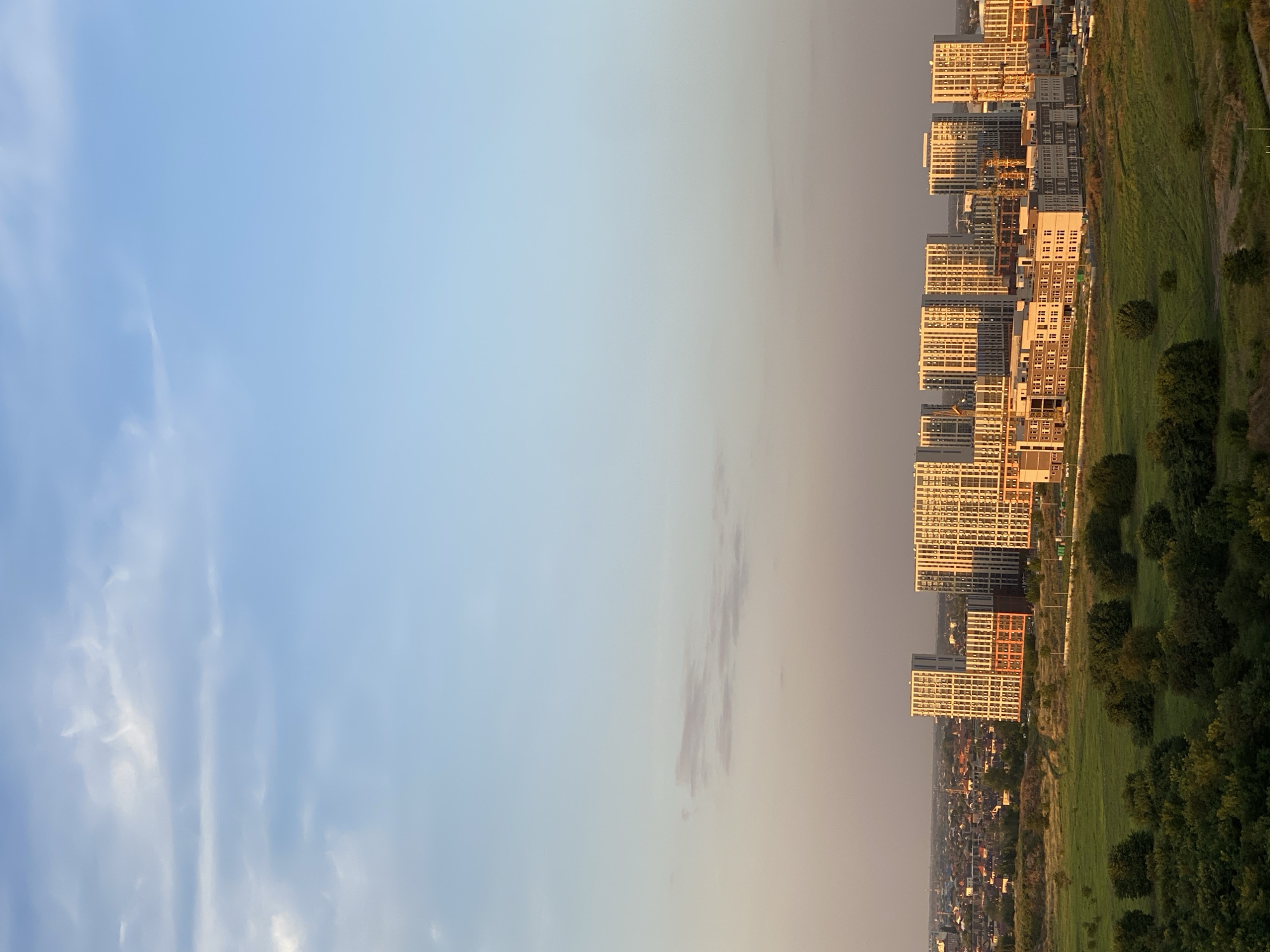 The image captures a late afternoon or early evening view of a developing urban area in Krasnodar, Russia, with modern high-rise apartment buildings dominating the skyline. The buildings are characterized by their light-colored facades and numerous windows, with some showcasing balconies. The setting sun casts a warm, golden light on the structures, highlighting their architectural details. In the foreground, a rolling green landscape with trees and grass suggests undeveloped or parkland areas surrounding the construction. A lower, older residential area with red-tiled roofs is visible in the middle ground to the left of the new buildings. The sky above is a gradient of soft blues and pastel tones, with scattered clouds, indicating a transition from day to evening. There are no people or vehicles directly visible in the frame. The overall scene depicts urban expansion and ongoing construction.