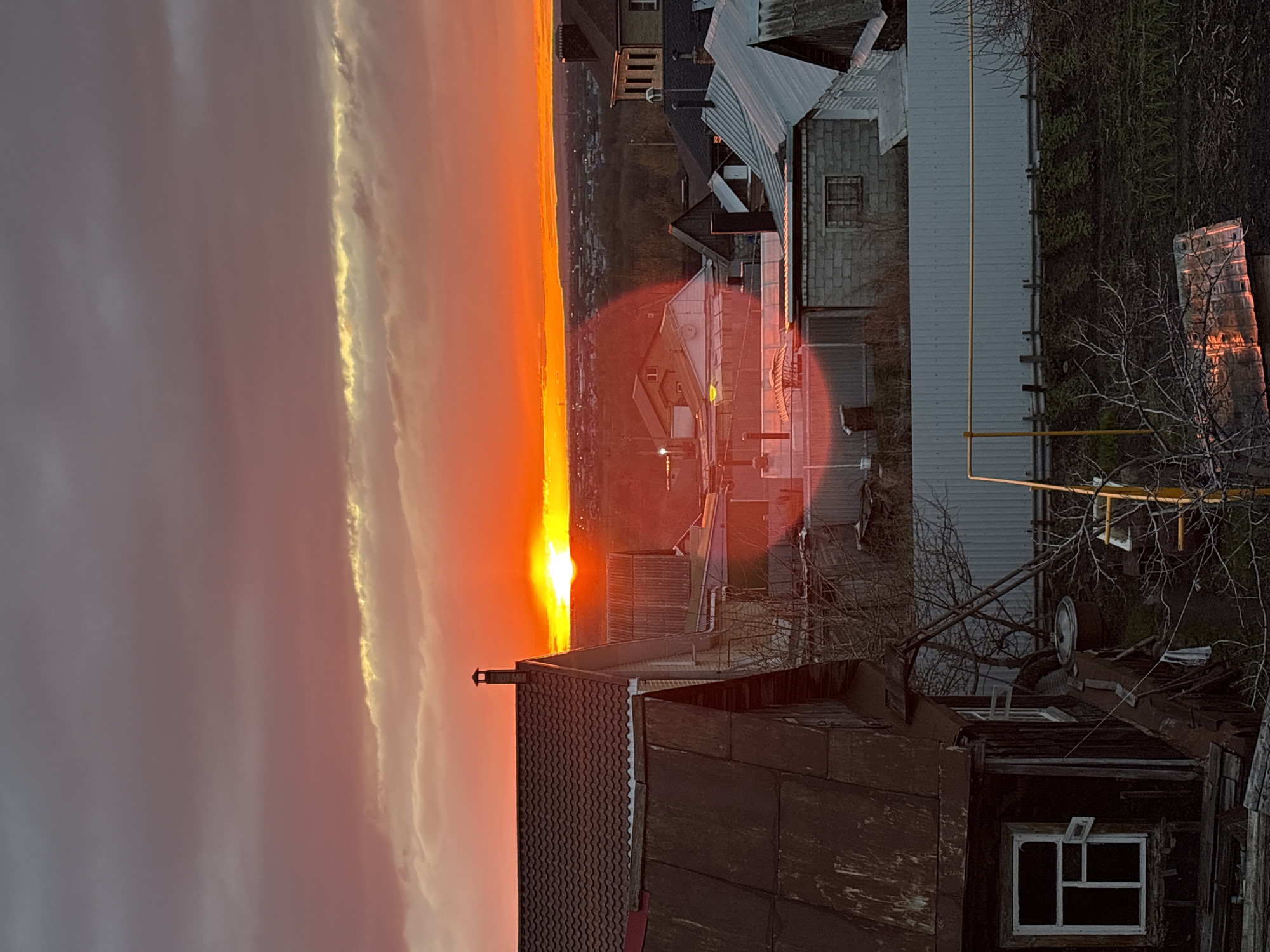 The image captures a vibrant sunset over a residential area in what is presumably Ulyanovsk, Russia, viewed from an elevated perspective. The time of day is clearly sunset, as an intense, glowing orb of the sun is visible low on the horizon, casting a dramatic orange and red hue across the sky.
The upper portion of the image is dominated by the sky, with the sun centered on the far left. A long, horizontal band of clouds stretches across the middle of the sky, its underside brilliantly illuminated in golden light by the setting sun, while the clouds above and below appear in shades of muted gray and purplish-pink. A notable red circular lens flare is present in the middle of the image, overlapping some of the buildings.
Below the horizon, a sprawling residential landscape unfolds. In the midground, numerous houses and buildings with various architectural styles are visible. Many roofs feature corrugated metal or tiling, and building materials range from brick to wood. A distinctive yellow gas pipe runs horizontally across the side of a building in the mid-right. Bare tree branches, devoid of leaves, are scattered throughout the scene, suggesting either late autumn or winter. In the foreground, in the bottom right corner, a section of a weathered, dark wooden structure with a white-framed window and a corrugated roof is prominently featured.
No people or specific activities are visible, rendering the scene tranquil. The overall impression is one of peaceful urban beauty bathed in the warm, dramatic light of dusk. No visible text is present in the image.