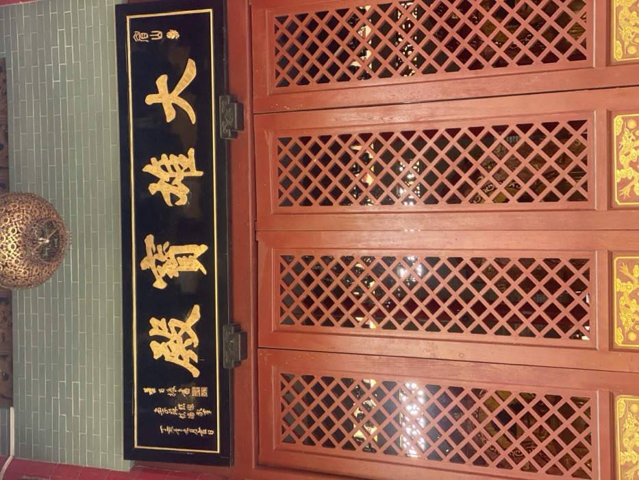 A vertical black plaque with prominent gold Chinese characters dominates the left side of the image, mounted on a reddish-brown wooden structure. The main characters on the plaque, read from top to bottom, are "大慈寶殿" (Dà Cí Bǎo Diàn), translating to "Great Compassion Treasure Hall." Above this, smaller gold characters read "南山" (Nán Shān), meaning "South Mountain." At the bottom of the plaque, even smaller characters indicate a dedication and a date, with the last visible line appearing to be "甲午年九月十八日" (Jiawu year, 18th day of the 9th month). A dark, possibly metal, hinge secures the plaque to the right.
To the right of the plaque are multiple horizontal panels of reddish-brown wood, featuring intricate diamond-shaped lattice patterns. These panels appear to be part of a larger door or wall structure, through which glimpses of an interior space with subtle reflections can be seen. On the far right, partially visible, are sections of vertical rectangular panels with ornate golden patterns, possibly depicting dragons or other auspicious symbols, against a red background.
On the far left, a decorative, spherical object with a complex, possibly metallic or ceramic, golden pattern hangs against a light green or grey tiled wall. No people are visible, and no specific activity is taking place. The scene depicts a static architectural detail, likely within a temple or traditional Chinese building in Guang Zhou Shi, China. The lighting suggests an indoor or sheltered outdoor setting.