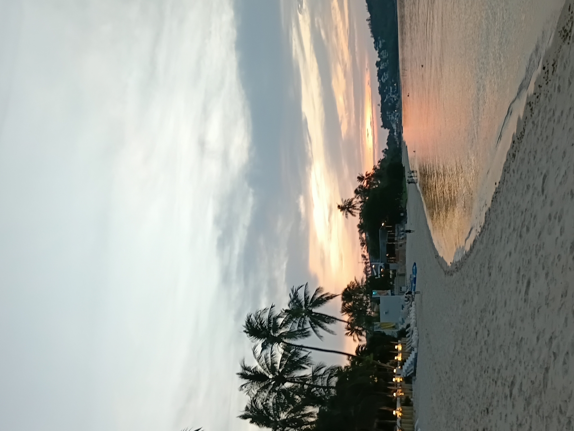 This image depicts a beach scene at sunset in Ko Samui District, Thailand. The sky is filled with soft clouds, tinged with orange and pink hues from the setting sun. Palm trees line the shore, their fronds silhouetted against the sky. The sand is a light beige, with a gentle curve leading towards the calm water. Several beach chairs are set up on the sand, some with people relaxing on them. In the distance, a few small buildings and resort-style structures are visible along the coastline, nestled amongst lush greenery. The water reflects the colors of the sunset, creating a serene and picturesque view. A few people are visible walking along the shoreline or wading in the shallow water. The overall impression is one of tranquility and natural beauty.
