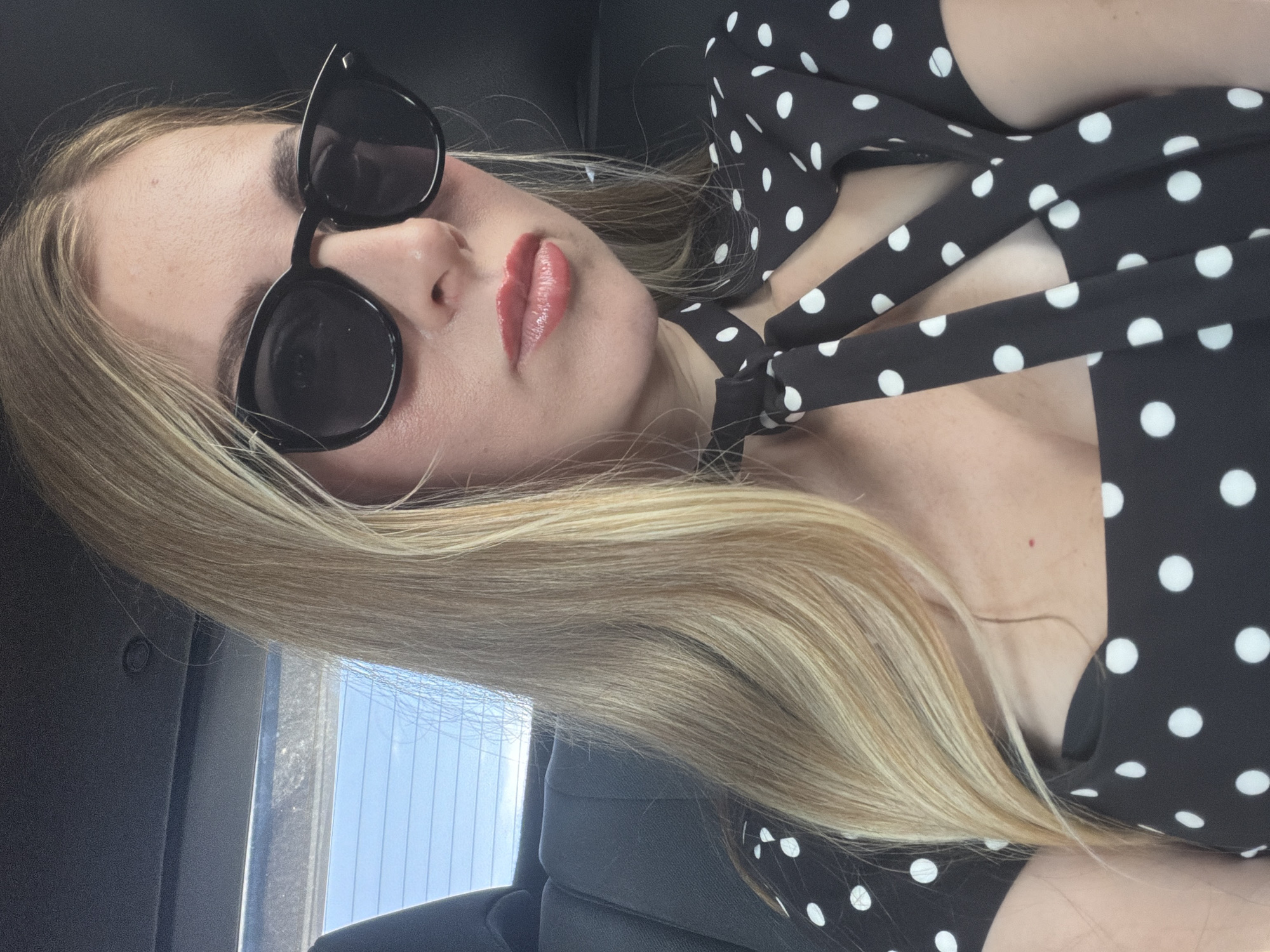 A woman with blonde hair and sunglasses is taking a selfie in the back seat of a car. She is wearing a black and white polka dot dress. The background shows the interior of a car, with the window behind her revealing a blue sky.