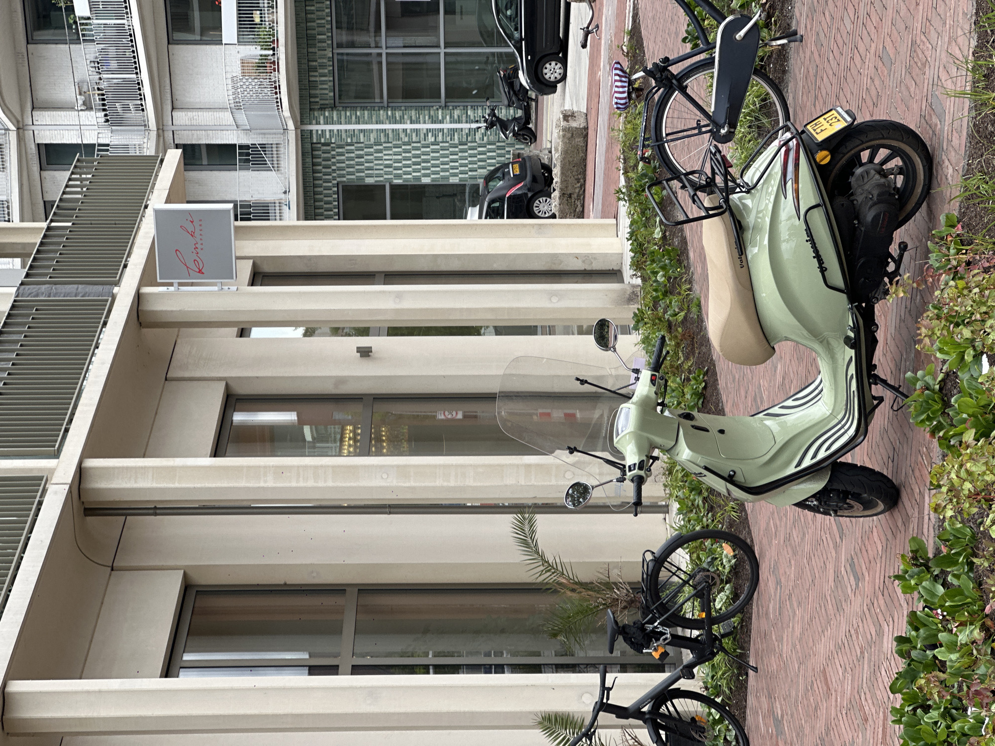 In Amsterdam, Netherlands, a light green Vespa scooter is parked on a brick walkway in front of a modern building with concrete pillars and large windows. A black bicycle is parked to the left of the scooter, and another black bicycle is partially visible to the right. In the background, a black car and a black motorcycle are parked on the street. A sign for "Rinki Kappers" is attached to the building above the scooter. Green plants are growing in front of the walkway.