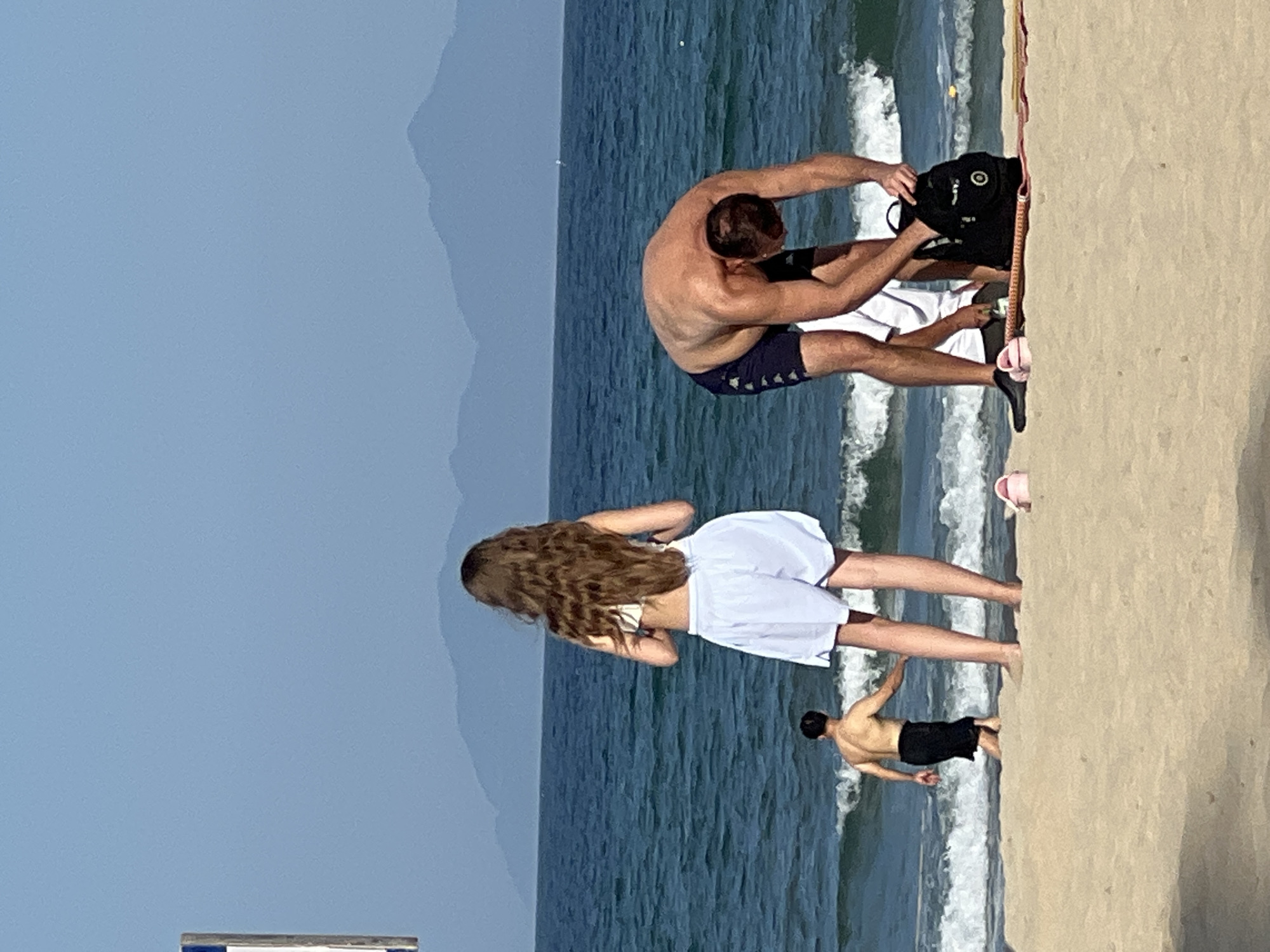 The image depicts a beach scene in Da Nang, Vietnam, on a clear, sunny day. The ocean is visible with moderate waves, and a hazy mountain range forms the distant horizon. In the foreground, a sandy beach is populated by several individuals. On the right, a man wearing dark blue swim trunks is bent over, retrieving items from a black backpack. To his left, a person in a white shirt is partially visible. Further left, a woman with long, wavy brown hair, wearing a white bikini top and white shorts, stands facing the ocean. Closer to the water, a man in black swim shorts is seen walking away from the camera towards the ocean. Two pink flip-flops are positioned on the sand in the middle ground. The scene conveys a relaxed atmosphere of a beach day.