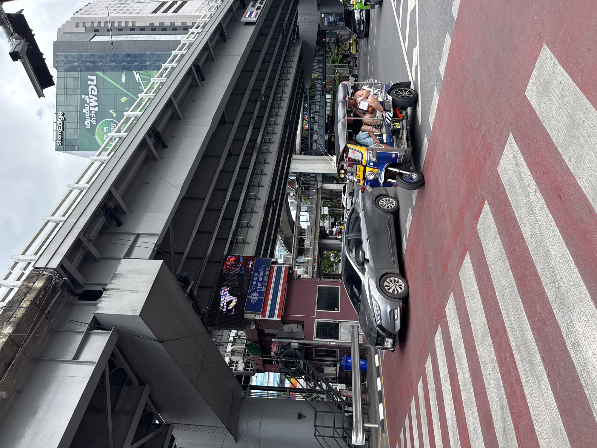 This image depicts a street scene in Bangkok, Thailand, likely during the daytime. In the foreground, a red and white striped pedestrian crossing is visible, with a gray sedan stopped on the left side of the frame and a blue and yellow tuk-tuk vehicle with three passengers in the center. The tuk-tuk appears to be in motion, navigating the intersection. Behind the vehicles, an elevated structure, possibly part of an expressway or bridge, dominates the upper portion of the image. A modern glass-fronted building with a green advertisement banner displaying Thai text and an image of elephants is visible on the right, suggesting a commercial or business district. To the left of the pedestrian crossing, a small, maroon booth with stairs leading up to it is situated under the elevated structure, with a Thai flag and a digital billboard displaying text and images above it. Traffic signals and other street infrastructure are also present in the background, indicating a busy urban environment. The sky is overcast, suggesting cloudy weather.