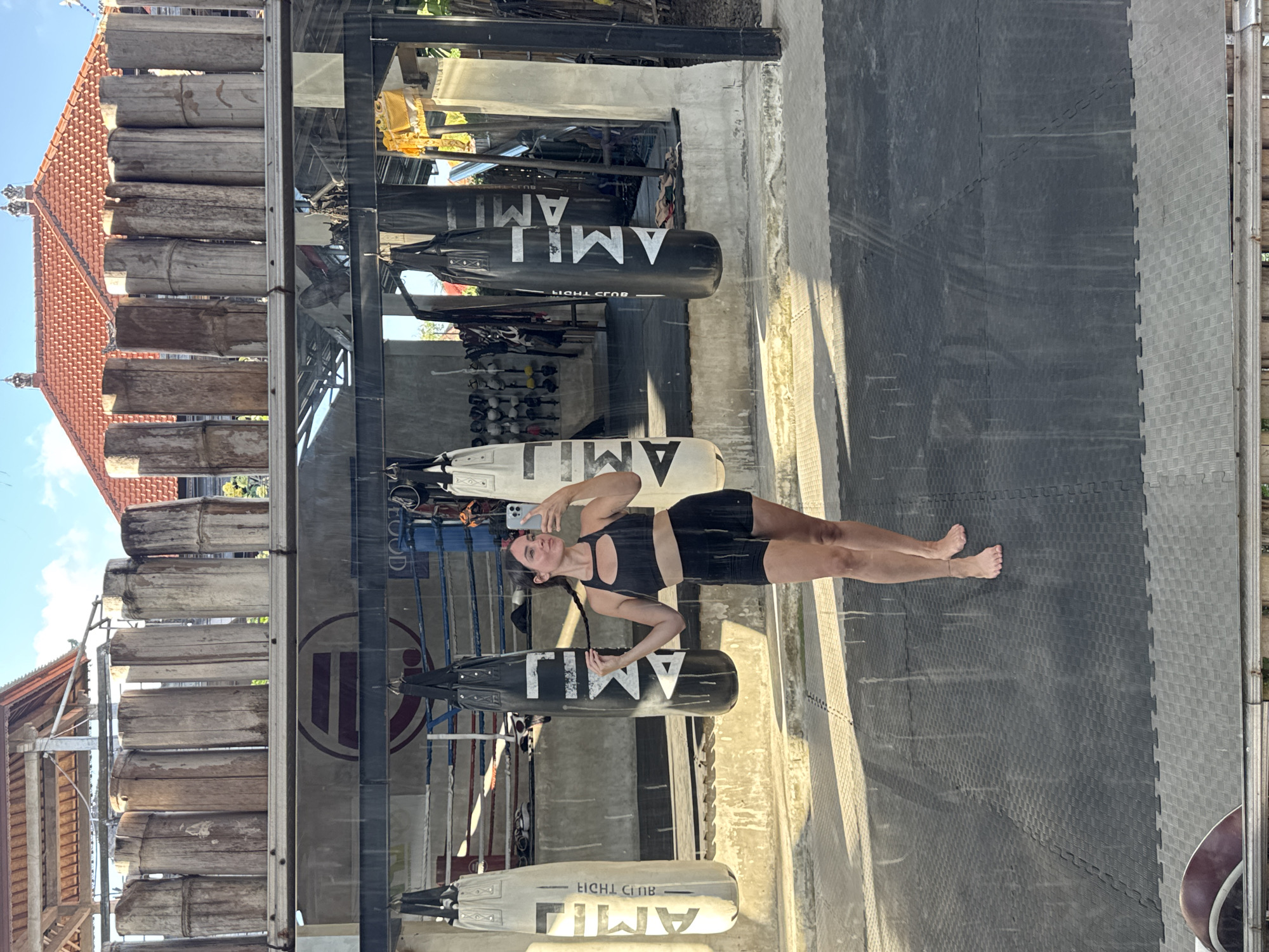 A woman takes a mirror selfie in a Muay Thai gym in Kuta Selatan, Indonesia. She is wearing a black sports bra and shorts and her hair is in a braid. She is standing in front of several punching bags, with a boxing ring in the background. The gym appears to be open-air, with a wooden roof and bamboo pillars. The floor is covered with black mats. The image is slightly blurry and there are water spots on the mirror, suggesting it may have been taken after a rain shower.