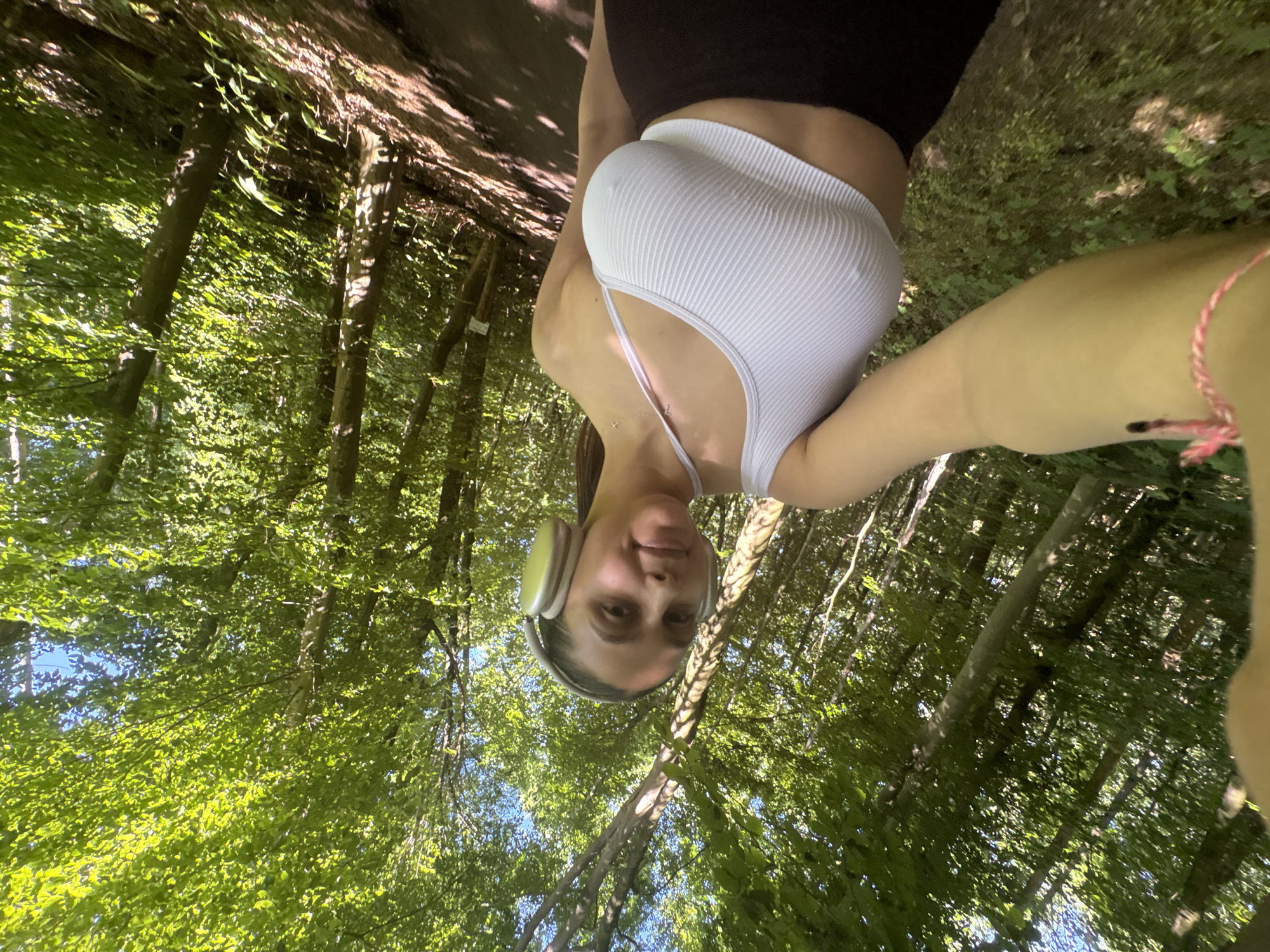 A young woman is taking a selfie in a wooded area. She is wearing a white ribbed tank top, black leggings, and headphones. She has a string bracelet on her wrist. The background shows trees with green leaves and a glimpse of a pathway. It is likely in the city of Viersen, Germany.