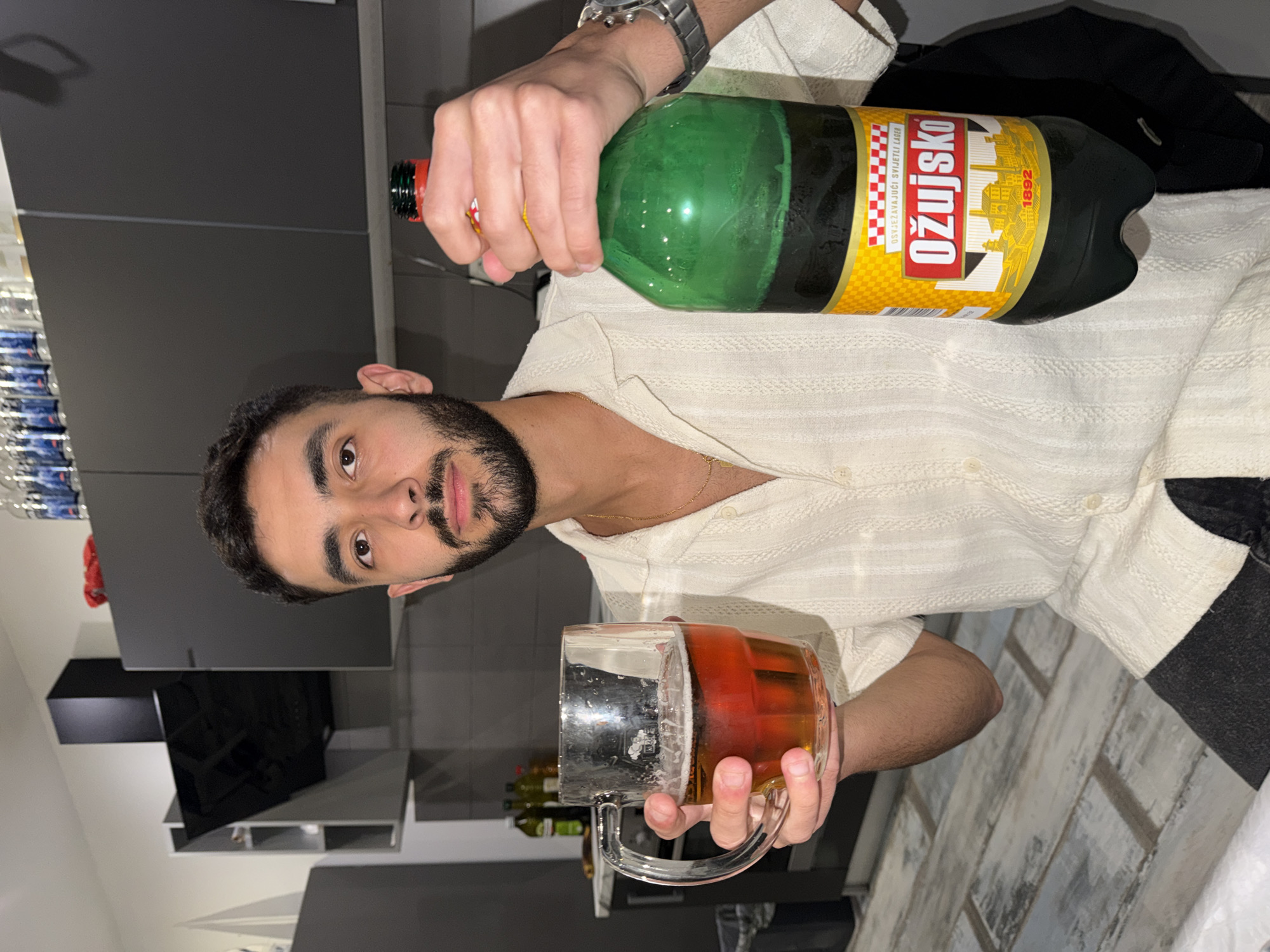 A young man with dark hair and a beard, wearing a light-colored, textured collared shirt and a gold chain, looks directly at the camera. He wears a watch on his left wrist. The image is taken indoors, likely in a kitchen or a home setting in Grad Rijeka, Croatia, identifiable by the prominent brand of beer.
In his right hand, he holds a large, green plastic bottle of "OŽUJSKO" beer, a Croatian brand, with a red cap. The yellow and red label clearly displays "OŽUJSKO" in bold red letters, along with the text "OSVJEŽAVAJUĆI SVIJETLI LAGER" (Refreshing light lager) and the year "1892", accompanied by an illustration of buildings. His left hand grasps a large, clear glass mug, partially filled with an amber liquid and a layer of foam, indicative of beer.
The background features modern, dark grey cabinetry or shelves, suggesting a contemporary interior. To the far left, a stack of bottled water is visible. A dark fabric item, possibly clothing, rests behind the beer bottle. The floor below appears to be light-colored, with a distressed or wood-grain pattern. The man's expression is neutral, with a slight hint of a smile, suggesting a casual and relaxed moment, likely enjoying a beverage.