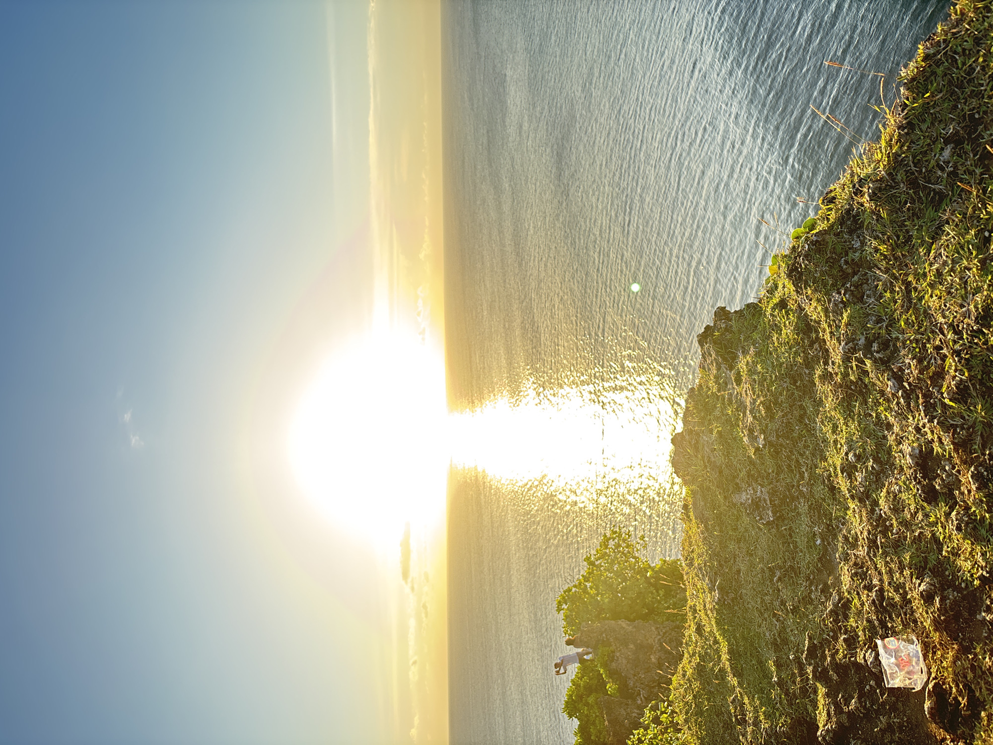 The image shows a landscape at Pecatu, Indonesia during sunset. The sun is setting over the ocean, creating a bright reflection on the water's surface. The sky is blue, fading to yellow near the horizon. The cliff is covered in green grass, a partially transparent plastic package lying on the grass. There is a person standing at the edge of the cliff, looking towards the ocean. The lighting suggests it is golden hour.