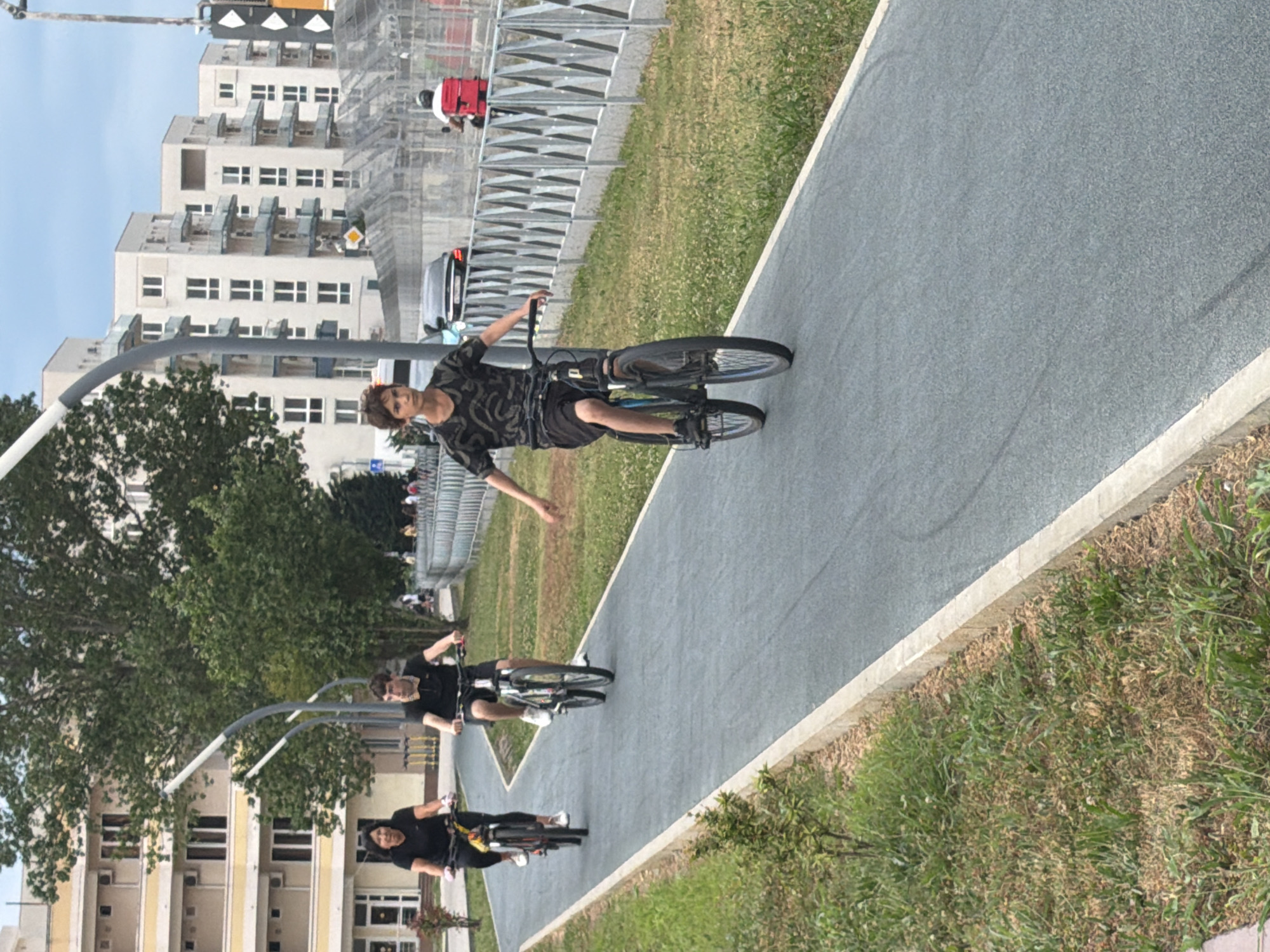 In Sirius, Russia, on a clear day, three individuals are cycling on a dedicated asphalt bike path. In the foreground, a young person with short brown hair, wearing a dark patterned t-shirt and black shorts, rides a dark bicycle with a blue frame. Their arms are outstretched, and they are looking forward. Behind them, slightly to the left, a person in a black t-shirt and dark pants rides a dark bicycle. To the left of that, a woman with dark hair, wearing a black t-shirt and white shoes, rides a yellow and black bicycle. The bike path is bordered by green grass and a concrete curb. In the background, there are modern apartment buildings with multiple balconies and windows. A metal fence runs along the right side of the bike path, and beyond that, there is traffic including a black car and a scooter with a red delivery box. The sky is partly cloudy.