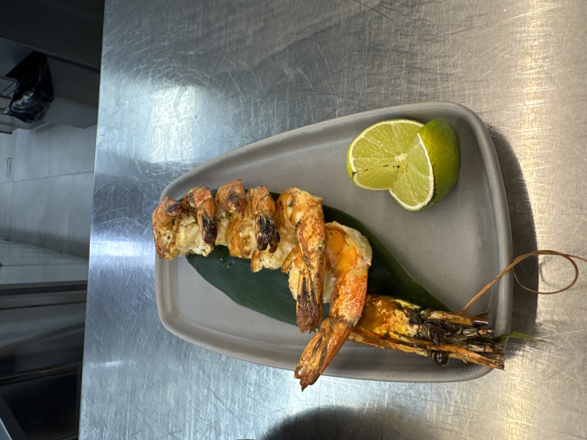 This image features a culinary dish, likely grilled shrimp, presented on a gray rectangular plate. Six to seven large, orange-red shrimp are arranged in a row, resting on a dark green leaf that spans the length of the plate. The shrimp appear cooked, with some exhibiting blackened grill marks and a creamy white seasoning or sauce. One shrimp on the far right retains its head and long, delicate antennae, which curl off the plate. Two bright green lime wedges, sliced in half, are placed on the upper right side of the plate.
The plate rests on a brushed metal surface, indicative of a professional kitchen countertop or prep area, with noticeable scratches and reflections. In the blurred background to the top left, a dark, crumpled object resembling a black plastic bag is visible, hanging against a light-colored wall. There is no visible text. The setting suggests a practical, utilitarian environment where food is prepared and plated.