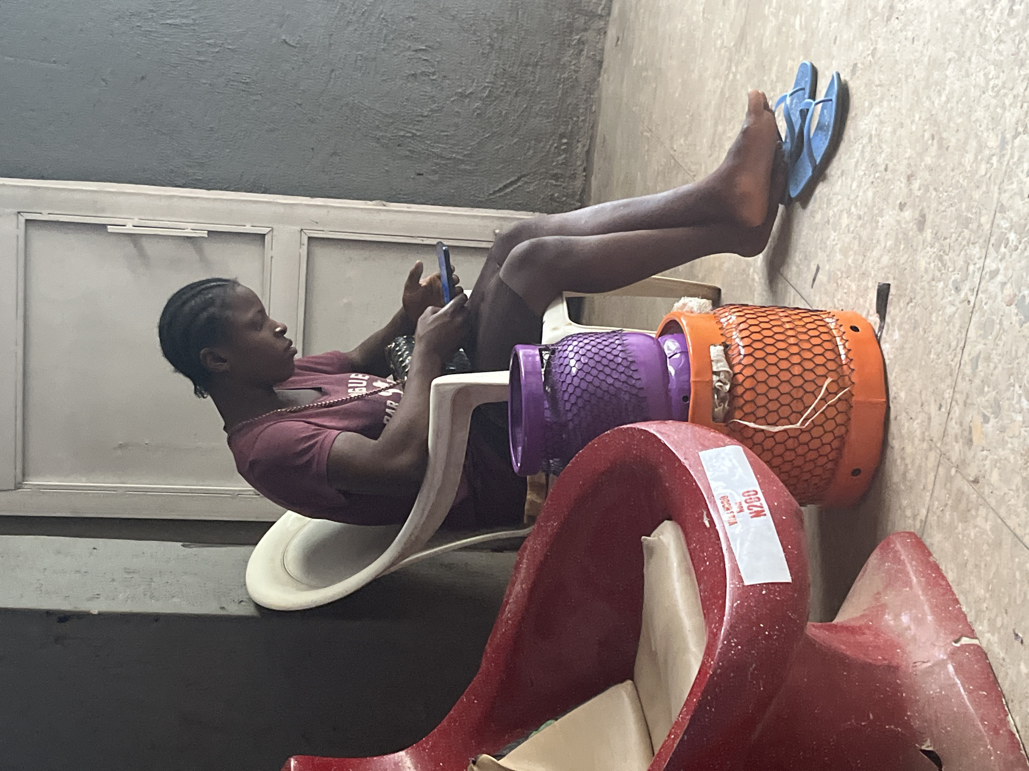 A young woman with dark skin and braided hair is seated in a white plastic chair, looking down at a smartphone in her hands. She is wearing a maroon t-shirt and dark bottoms. To her left, in the foreground, is a red, oddly shaped plastic object that appears to be a type of seat or basin with a light-colored cushion. Next to the chair, and partially behind the red object, are two stacked cylindrical containers, the bottom one orange with a mesh pattern and the top one purple with a similar mesh pattern. A pair of blue flip-flops lies on the floor near the woman's bare feet. The background consists of a textured gray wall and a white door. The floor is tiled with a light-colored, speckled pattern. The scene appears to be an indoor waiting area, possibly at a public facility or shop, in Jos, Nigeria.