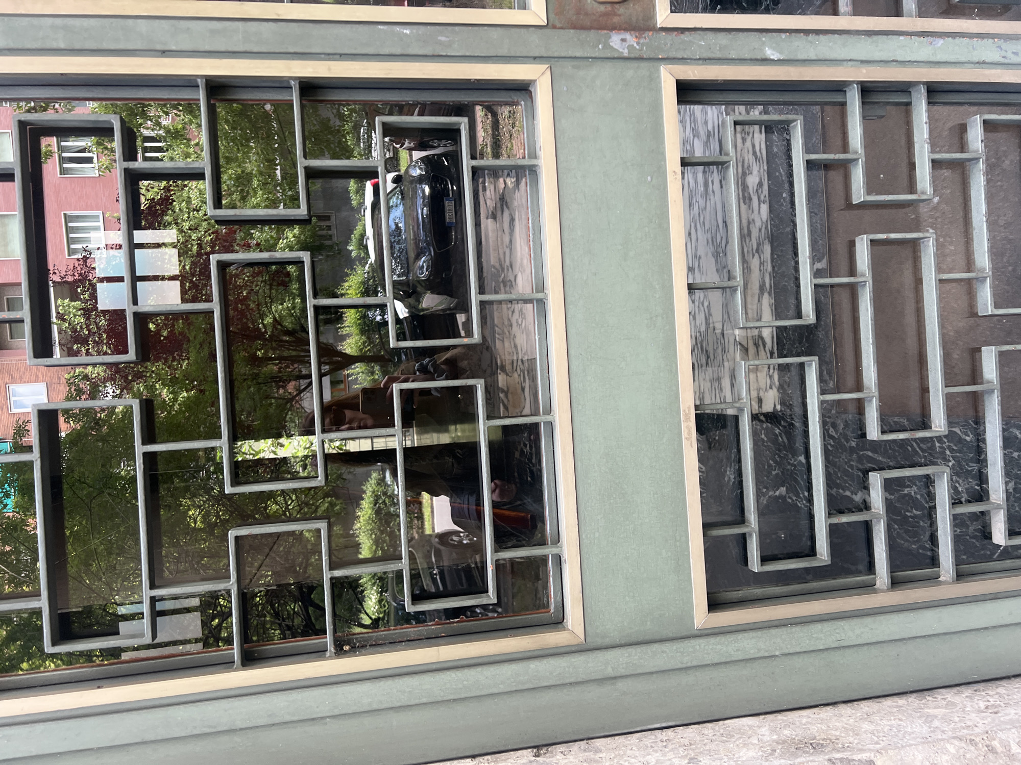 A close-up of an exterior door in Milan, Italy, featuring a distinctive geometric metal grille design over glass panels. The left panel reflects a street scene with trees, a building with salmon-colored walls, and a parked black car. The right panel shows a dark, marble-like background. The door frame is painted a muted green. The door is located on a street, as indicated by the visible sidewalk. The geometric grille features arrangements of rectangles and squares. A person is reflected in the glass.