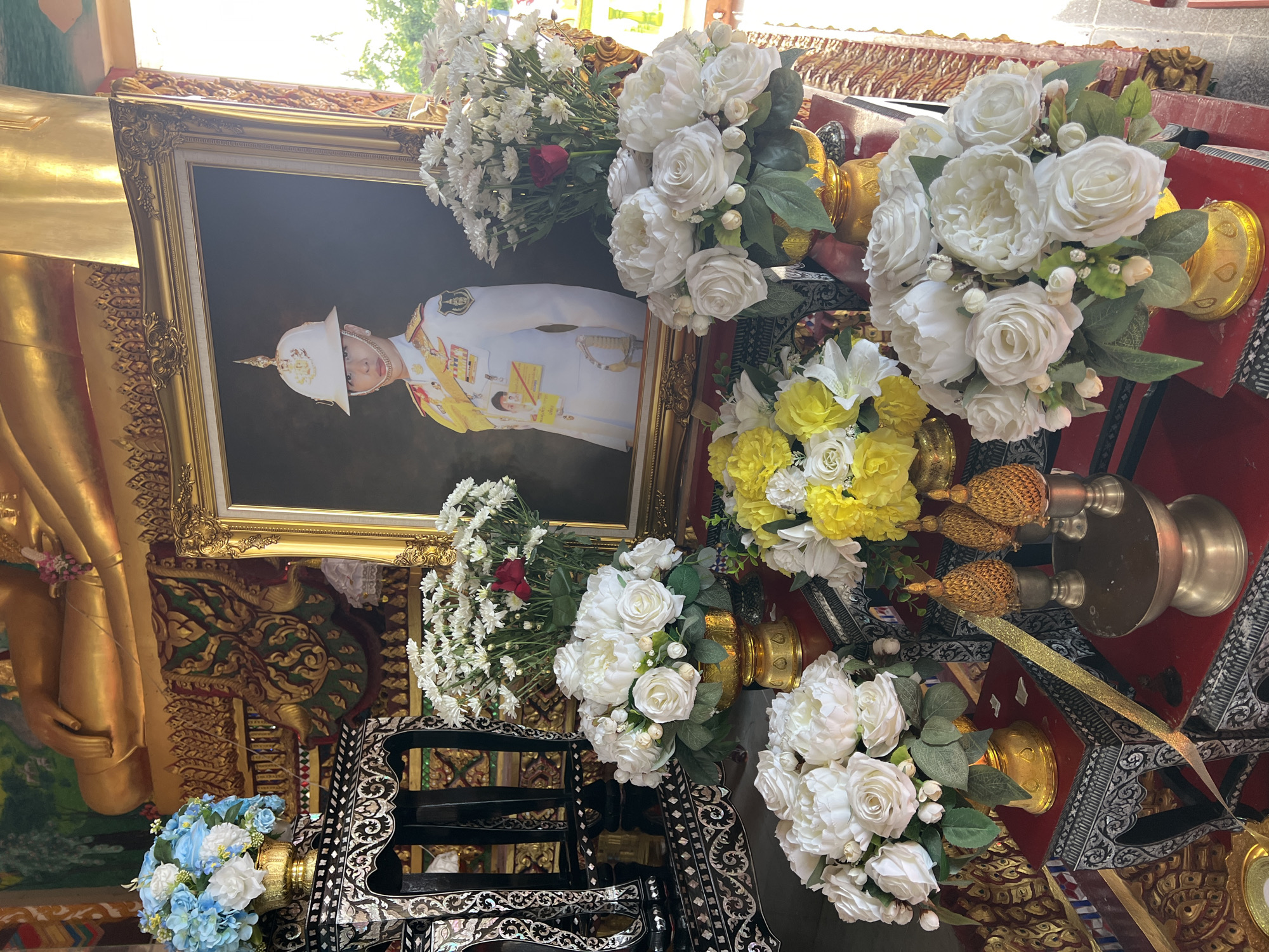 The image depicts an indoor shrine or altar in Baan Plai Laem, Thailand, likely within a Buddhist temple given the presence of a large golden Buddha statue in the background. The focal point of the arrangement is a framed portrait of a person wearing a white military uniform and a distinctive helmet, positioned centrally on the altar. Surrounding the portrait are numerous bouquets of artificial flowers. These include large white roses in several arrangements, a bouquet of white and blue flowers, and a cluster of yellow and white flowers. The altar itself is adorned with ornate golden containers for the flowers and decorative golden tassels. Behind the altar, the base of the golden Buddha statue is visible, with intricate golden decorative patterns. The overall impression is one of reverence and ceremonial display.
