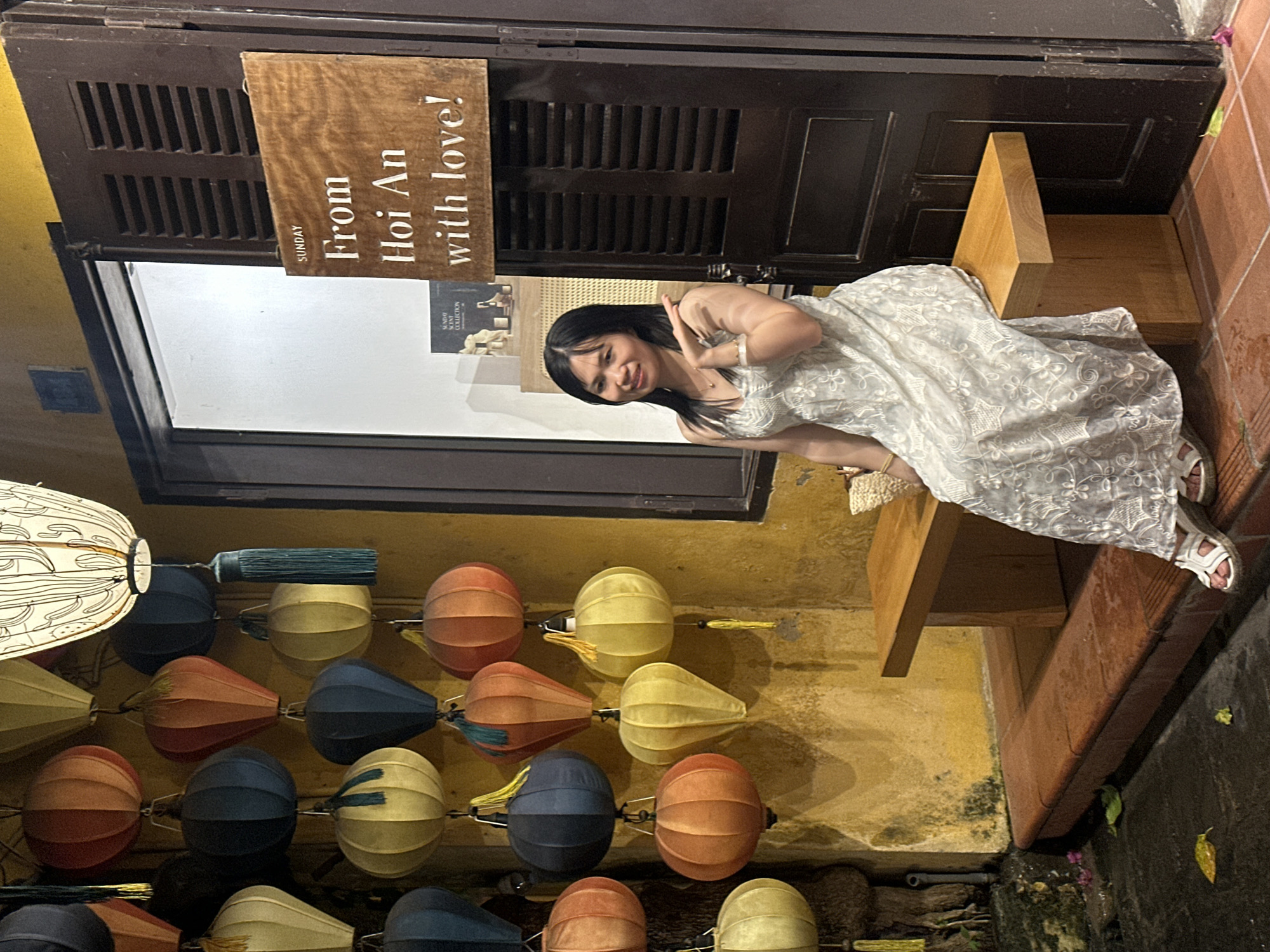 In a nighttime scene, a young woman in a white lace dress sits on a wooden bench outside a shop. She is smiling and making a peace sign with her fingers. Behind her, a dark wooden door with louvers is visible, with a brown sign hanging from it that reads "SUNDAY From Hoi An with love!". Below the door, a row of colorful, round paper lanterns hangs, illuminated by an unseen light source, creating a warm and inviting ambiance. The lanterns are a mix of yellow, orange, blue, and red, with some featuring decorative tassels. The wall behind the lanterns is a textured yellow, suggesting an outdoor setting in a location with traditional architecture. The ground appears to be dark and wet, possibly indicating recent rain. The overall mood is cheerful and relaxed, capturing a moment of enjoyment during a trip to Hội An.