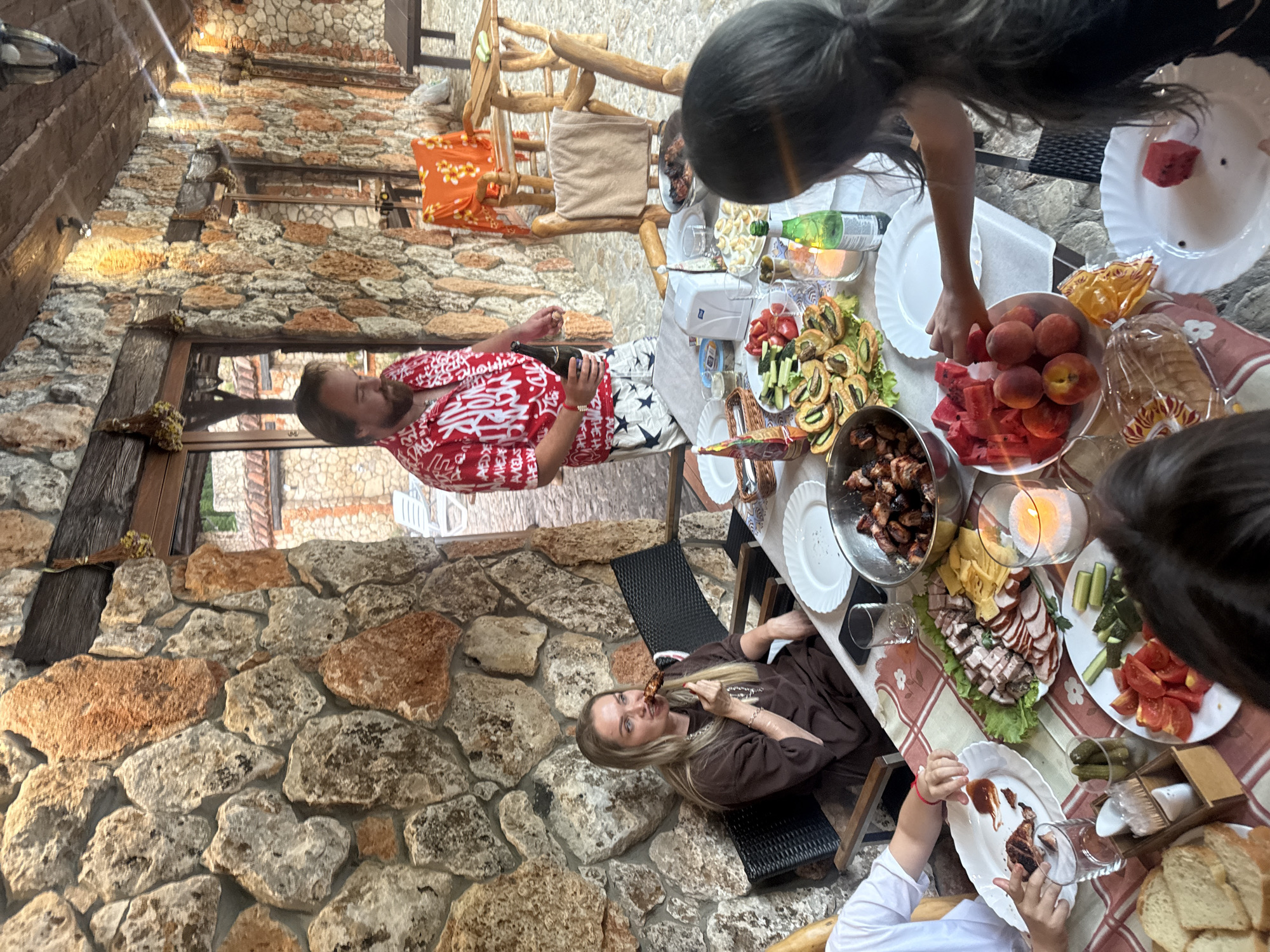 The image depicts an outdoor gathering at a stone-built structure, likely an extension or patio of a house, within the city of Krasnomakskoye rural settlement, Ukraine. A table laden with food, including grilled meat, sliced meats and cheese, vegetables, sliced fruits (peaches and watermelon), and bread, is central to the scene. Several people are present, including a woman in a brown sweatshirt eating from a skewer, a man in a red patterned shirt holding a bottle, and a child with long dark hair reaching for fruit. The environment features stone walls, wooden beams, and outdoor seating. The time appears to be daytime, with natural and artificial lighting present.