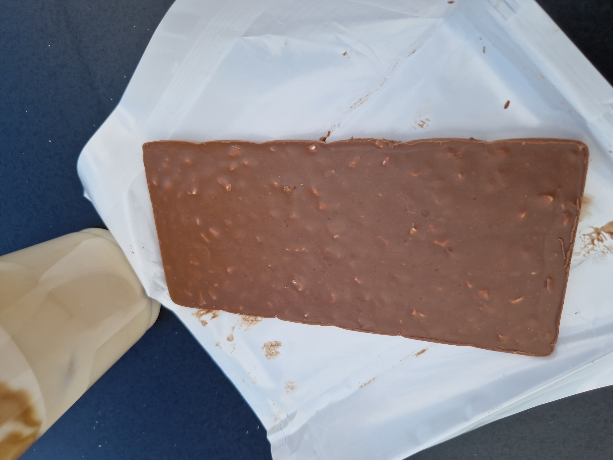 A rectangular chocolate bar with visible nuts is placed on a white paper wrapper. A clear plastic cup filled with a pale liquid, possibly coffee with milk, is positioned to the upper left of the chocolate bar. The scene is set on a dark, textured surface, likely a table or outdoor surface. The image was taken in Sofia, Bulgaria.