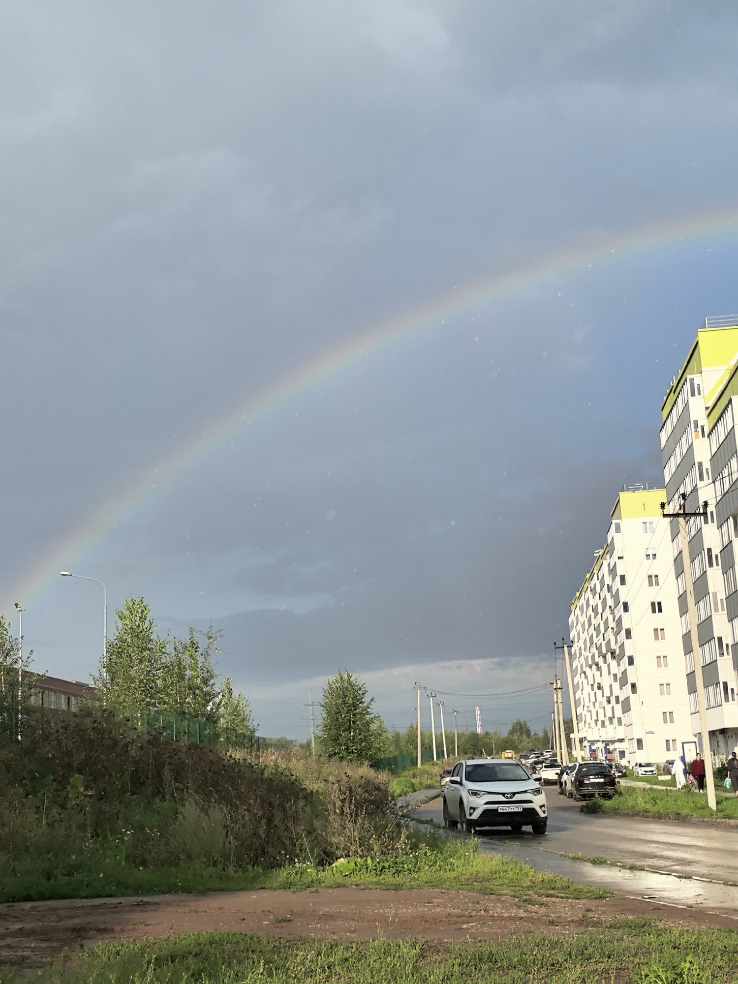 The image depicts a street scene in Froli, Russia, under a cloudy sky with a prominent rainbow arcing across it. In the foreground, a dirt road with patches of green grass is visible. To the right, a modern, multi-story apartment building with white and yellow facade stands tall. A paved road runs alongside the building, with several cars parked and driving on it. A white SUV is in the left lane of the paved road, facing away from the viewer. Several people are walking on the sidewalk near the building. Trees and foliage line the left side of the road, with a fence and another building in the distance. Electrical poles and wires are scattered throughout the scene, indicating an urban or suburban environment. The sky is a mix of dark clouds and lighter areas, with the rainbow being a vibrant focal point.