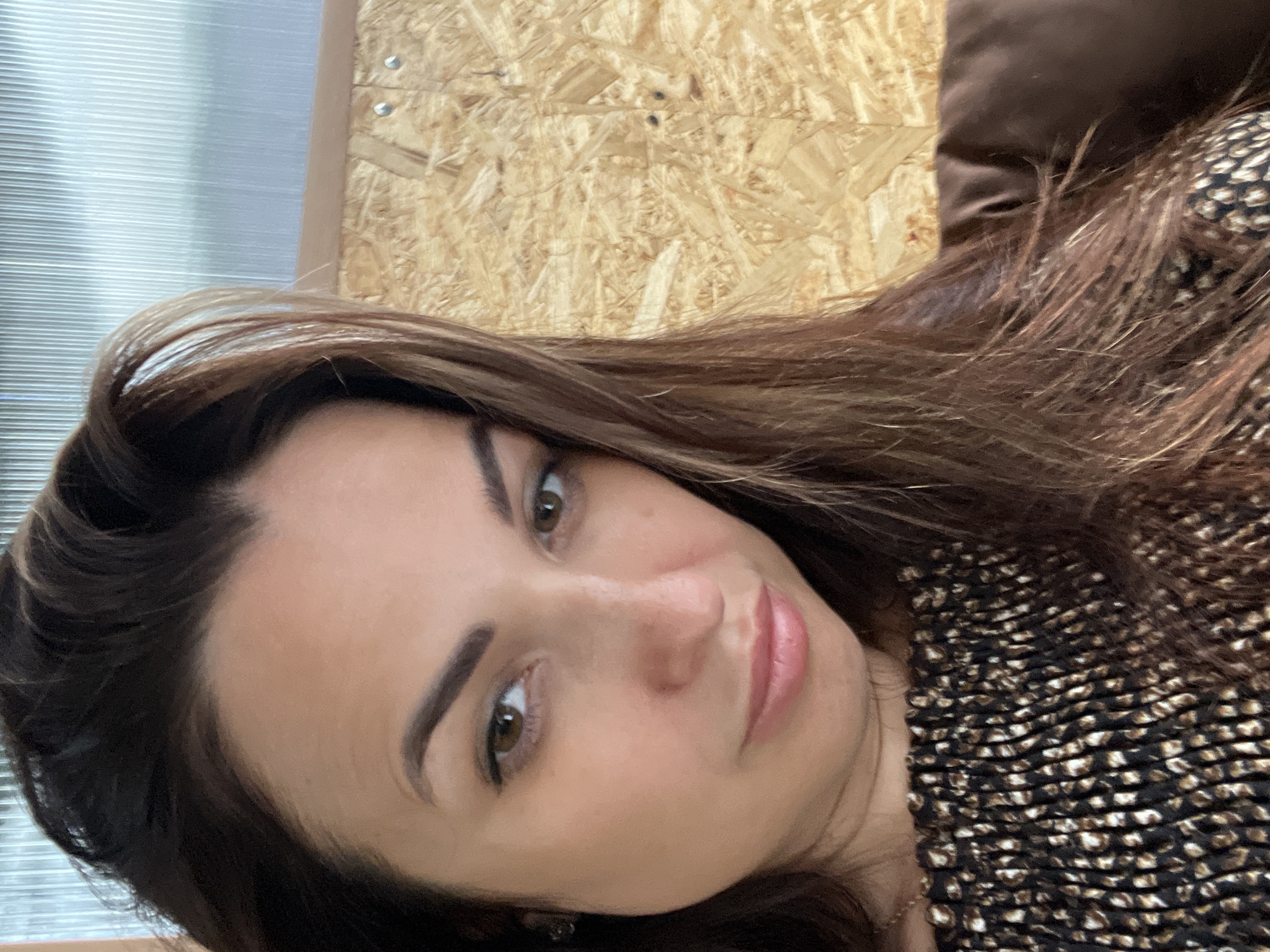 The image is a close-up, portrait-style photograph of a woman. She has long, dark brown hair that falls around her face and shoulders. Her eyes are brown and she is looking towards the camera. Her lips are slightly parted and appear to be painted with a light pink lipstick. She is wearing a dark-colored top with a patterned design.
The background of the image is blurred but appears to be an indoor setting. A textured, light-colored wall is visible behind her, possibly made of wood panels. To the left, there is a suggestion of blinds or a window. The lighting suggests it could be daytime, with natural light illuminating her face. The overall mood is relaxed and casual. There is no visible text in the image.