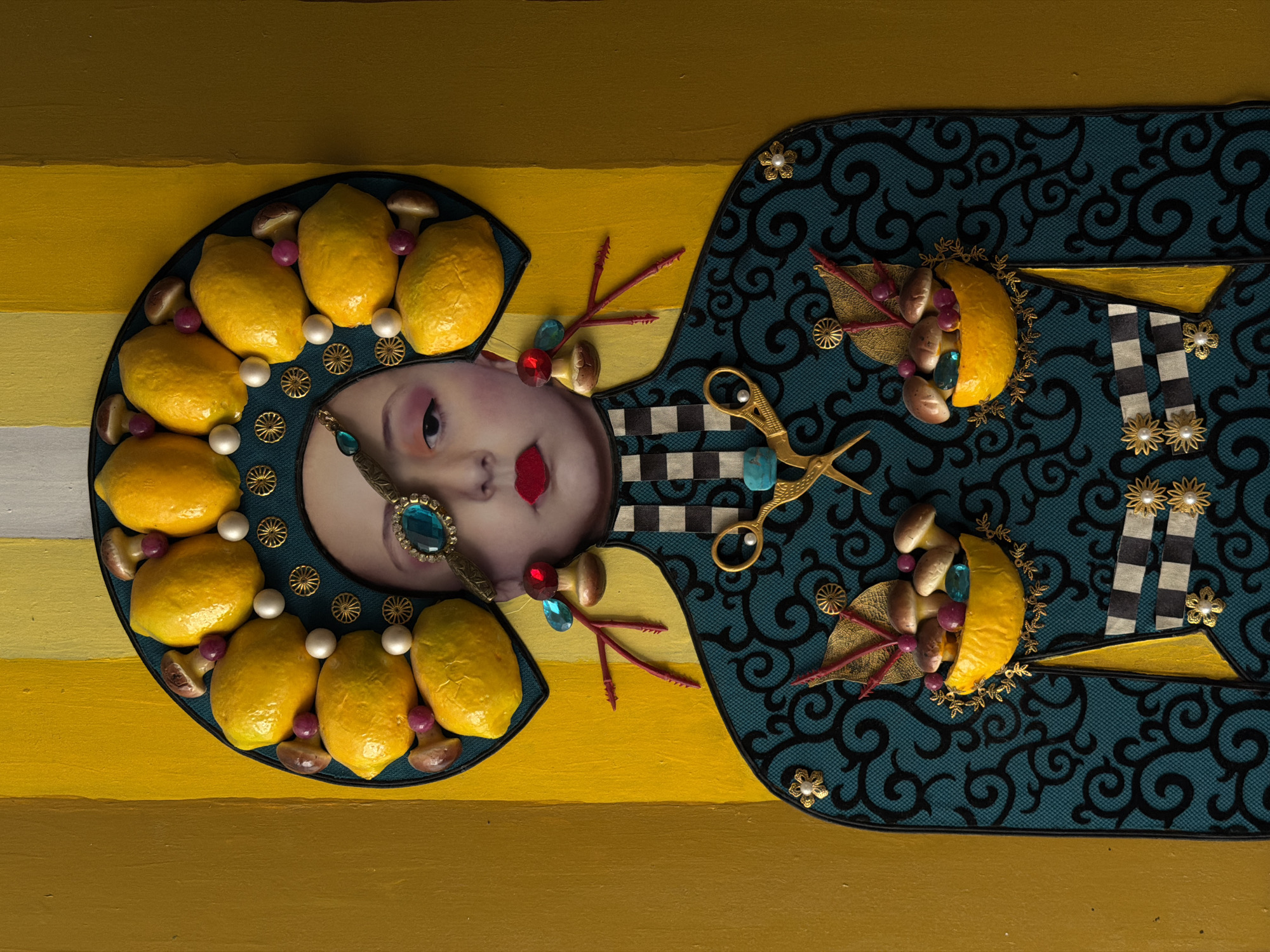 The image depicts an artistic assemblage on a yellow striped background. A centrally positioned face with red lips and a bejeweled monocle is surrounded by a headdress composed of lemons, mushrooms, and pearls. The figure also features an ornate, blue patterned garment adorned with lemons, mushrooms, scissors, and decorative flowers. The location in Yerevan, Armenia is not discernible from this image.
