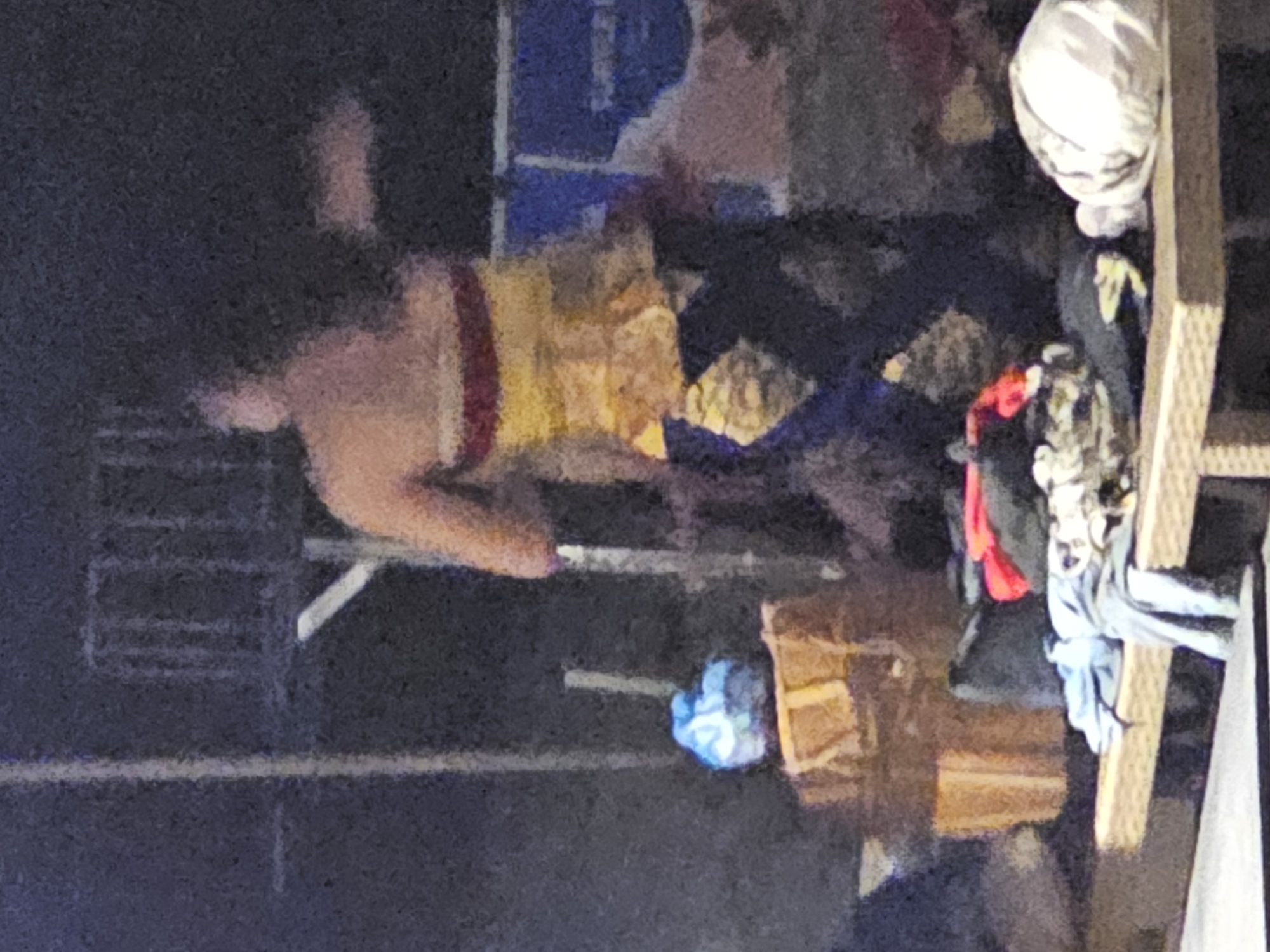A dimly lit scene depicts a person standing indoors. They are wearing a top that is striped red and yellow. There is a blue item near a wooden box. A rack with clothing items is also present. The location appears to be cluttered with miscellaneous objects.