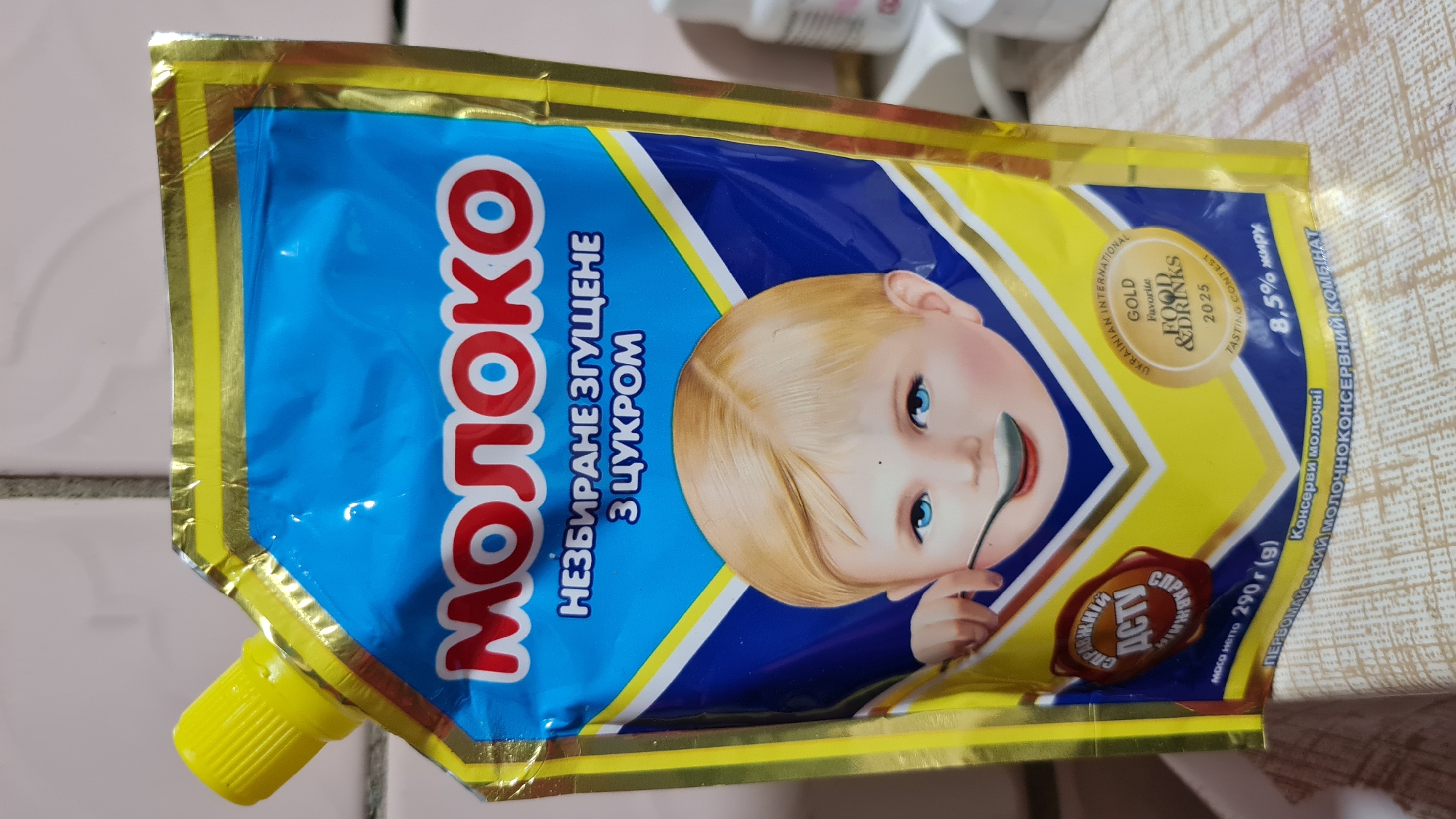 The image displays a close-up view of a pouch of "Молоко" (Milk), a Ukrainian brand. The pouch features a blue background with gold trim and depicts a young child with bright blue eyes, holding a spoon. Text on the pouch indicates it is "НЕЗБИРАНЕ ЗГУЩЕНЕ З ЦУКРОМ" (Unprocessed Condensed with Sugar). A seal with the text "СПРАВЖНІЙ ДСТУ" (Genuine DSTU) is also visible. Towards the bottom, it states "МОЛОКО НЕЗБИРАНЕ 290 g (g)" and "ПЕРВОМАЙСЬКИЙ МОЛОЧНОКОНСЕРВНИЙ КОМБІНАТ" (Pervomaisky Dairy Canning Combine). A gold award seal reads "UKRAINIAN INTERNATIONAL GOLD Favorite FOOD & DRINKS 2025 TASTING CONTEST" and "8,5% жиру" (8.5% fat). The pouch is resting on a surface that appears to be a tablecloth with a patterned texture. The overall scene is a product photograph, likely taken indoors, with no specific identifiable urban features of Salhany, Ukraine, present in the frame.