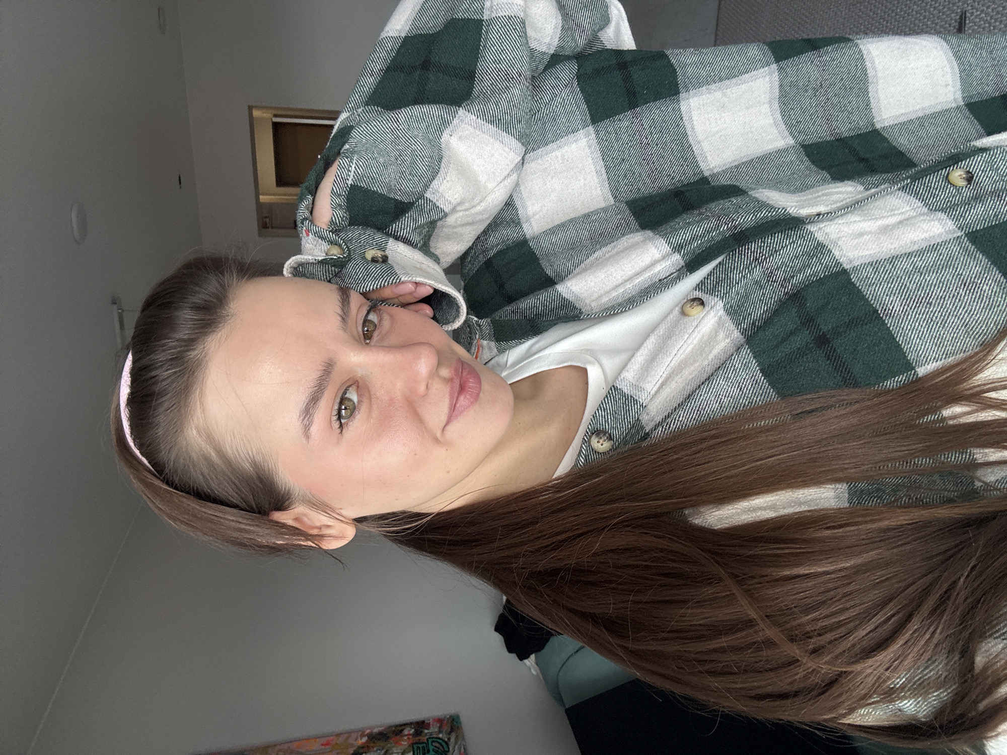 A woman with long brown hair is posing for a selfie. She is wearing a white t-shirt and a green and white plaid flannel shirt. She has a pink headband on. The background is a grey wall and a window. The city of Krasnoyarsk, Russia is not visible and cannot be determined from the image.